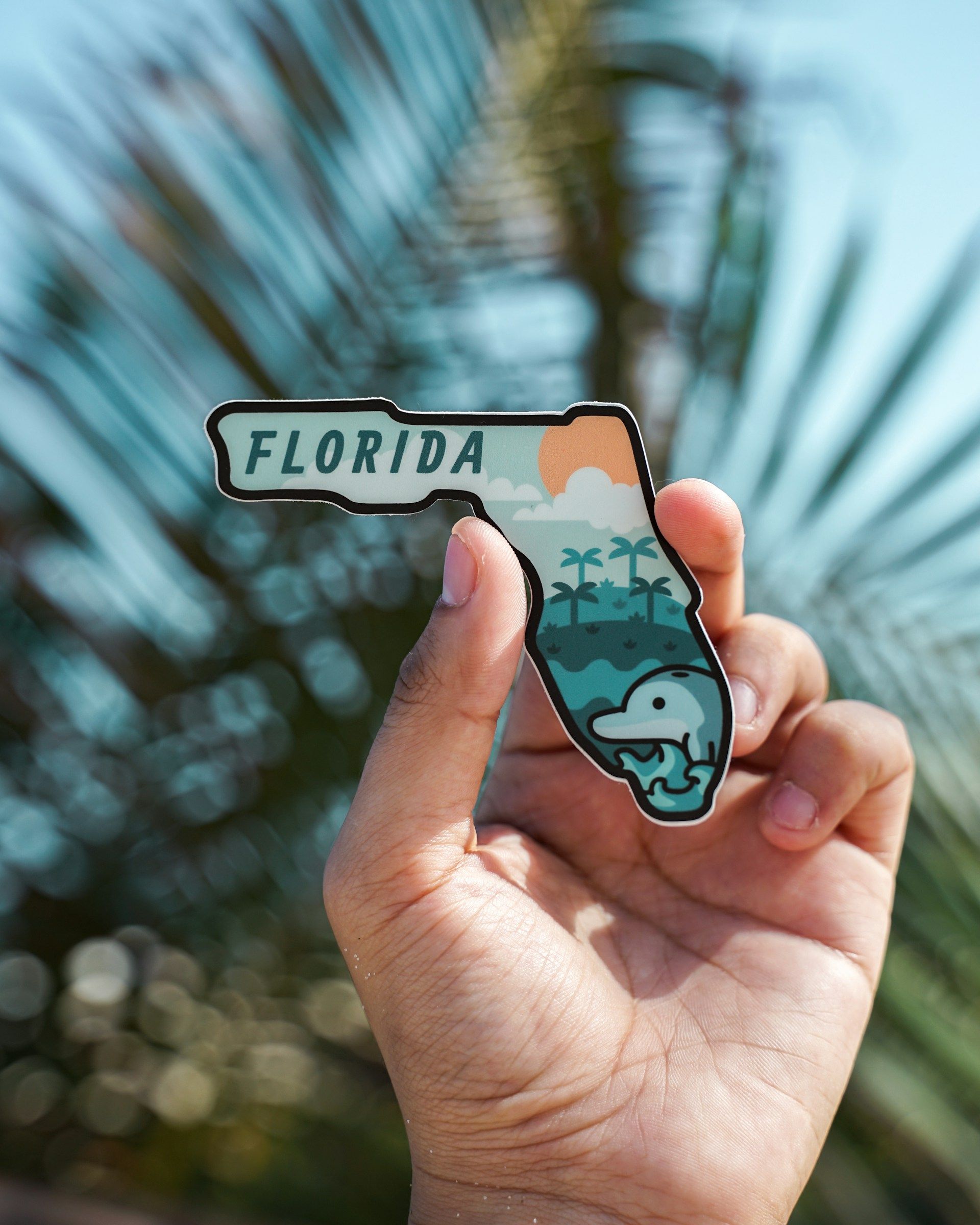 Hand holding a Florida-shaped pin with illustrations of a beach, palm trees, and an alligator.