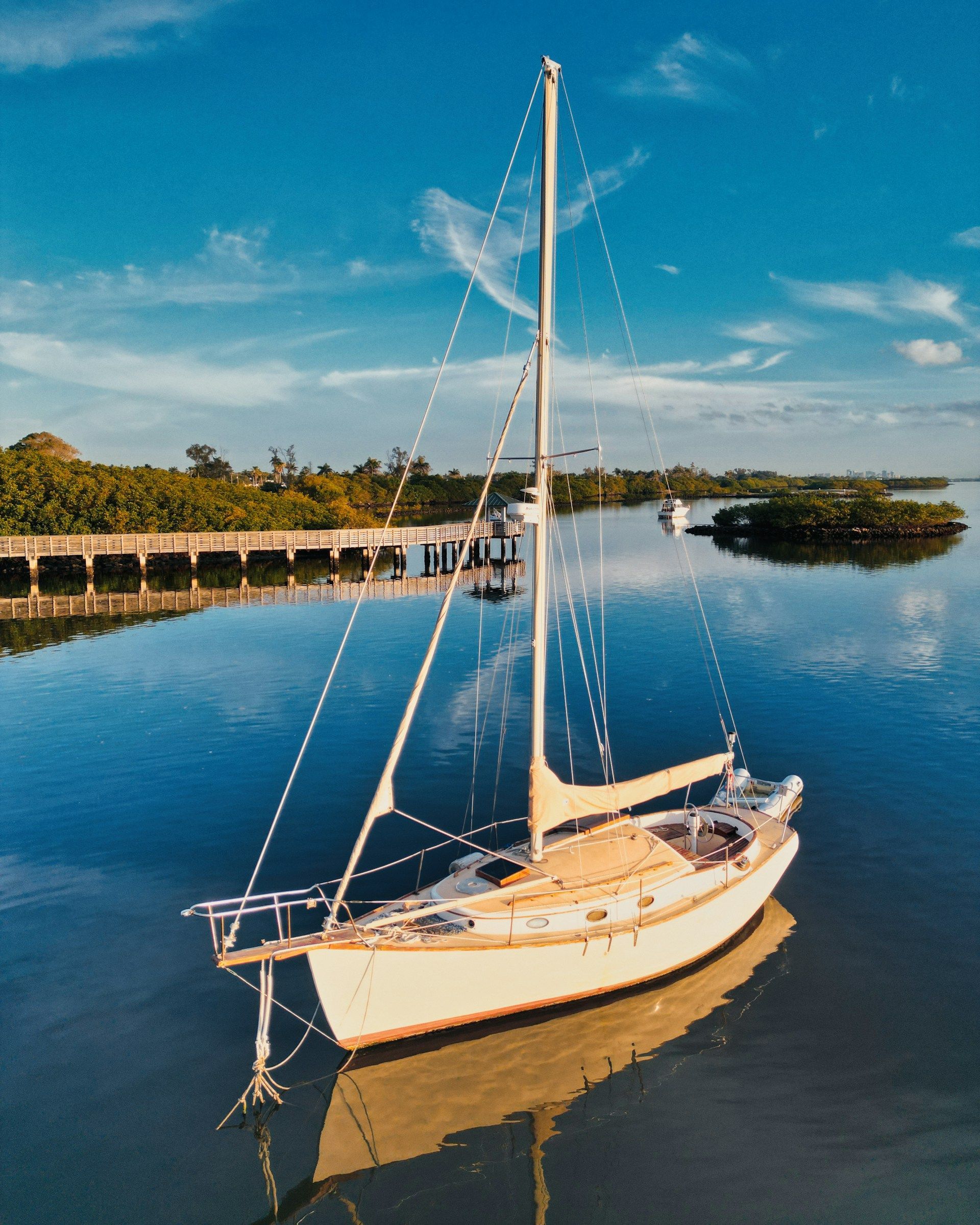 Sailboat on calm water; pier and mangrove trees in background under a blue sky.