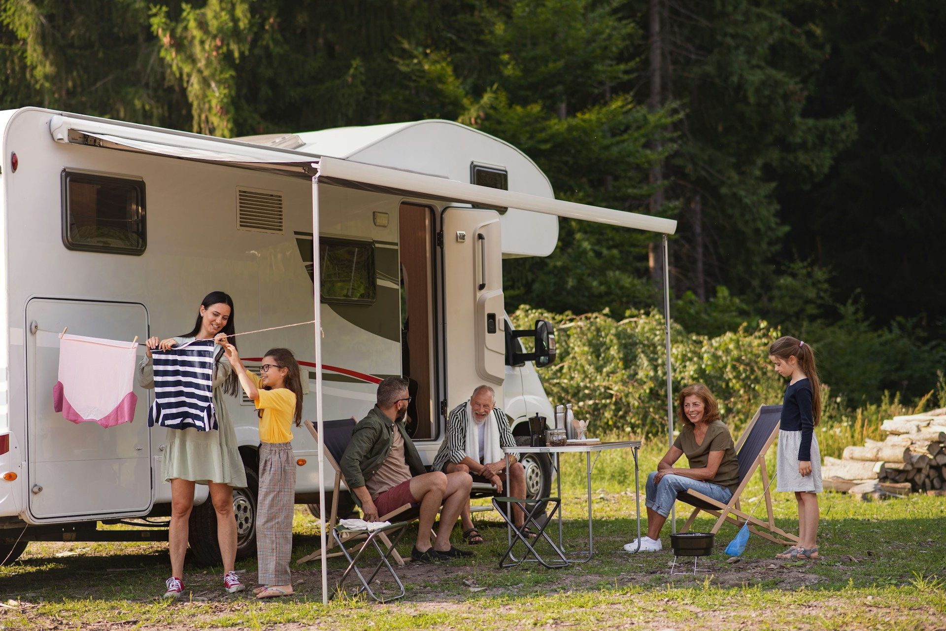 Family camping: woman and girl hang clothes near RV; others relax in chairs.