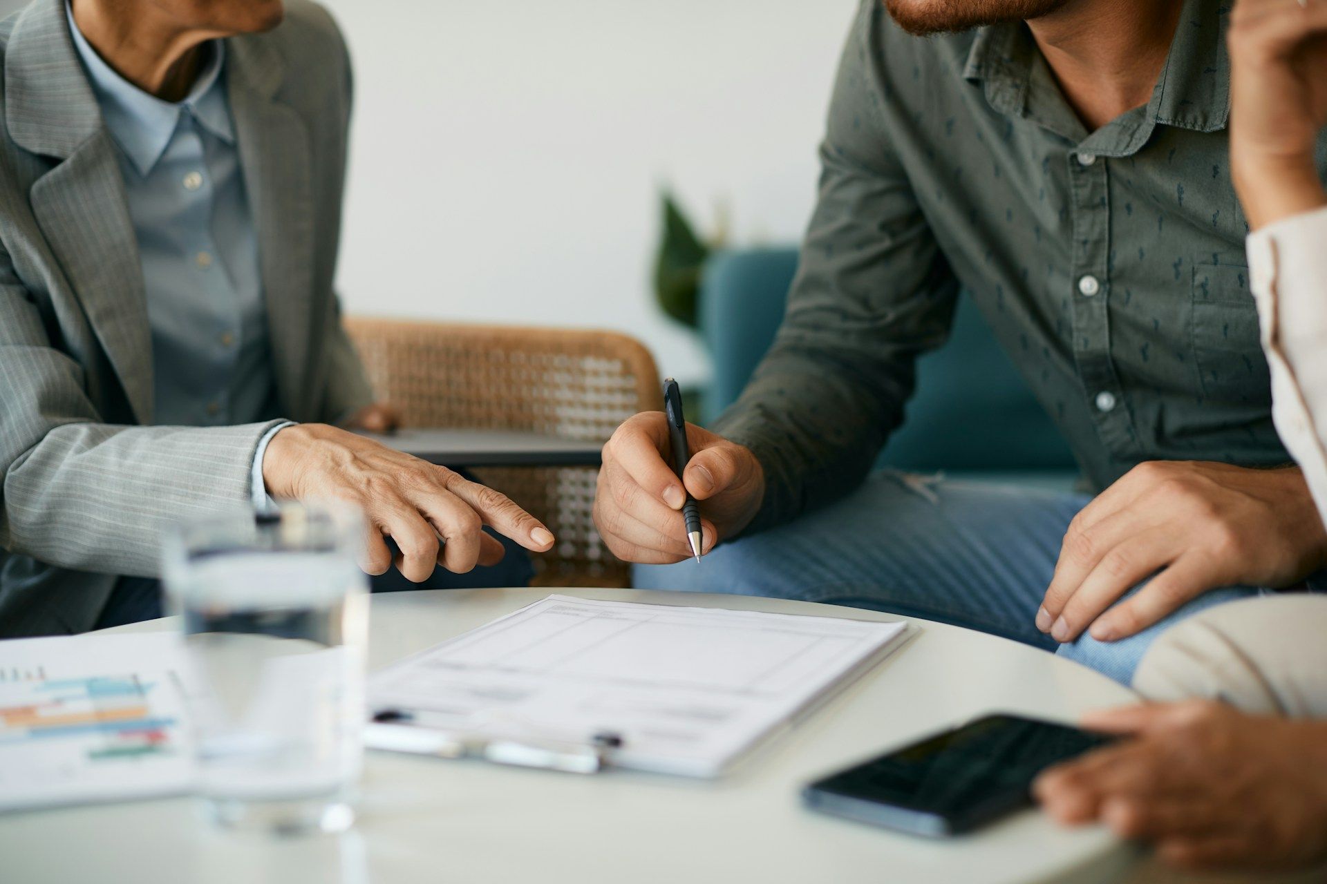 Person signing a document on a table; another person points to it. Glass of water, phone visible.
