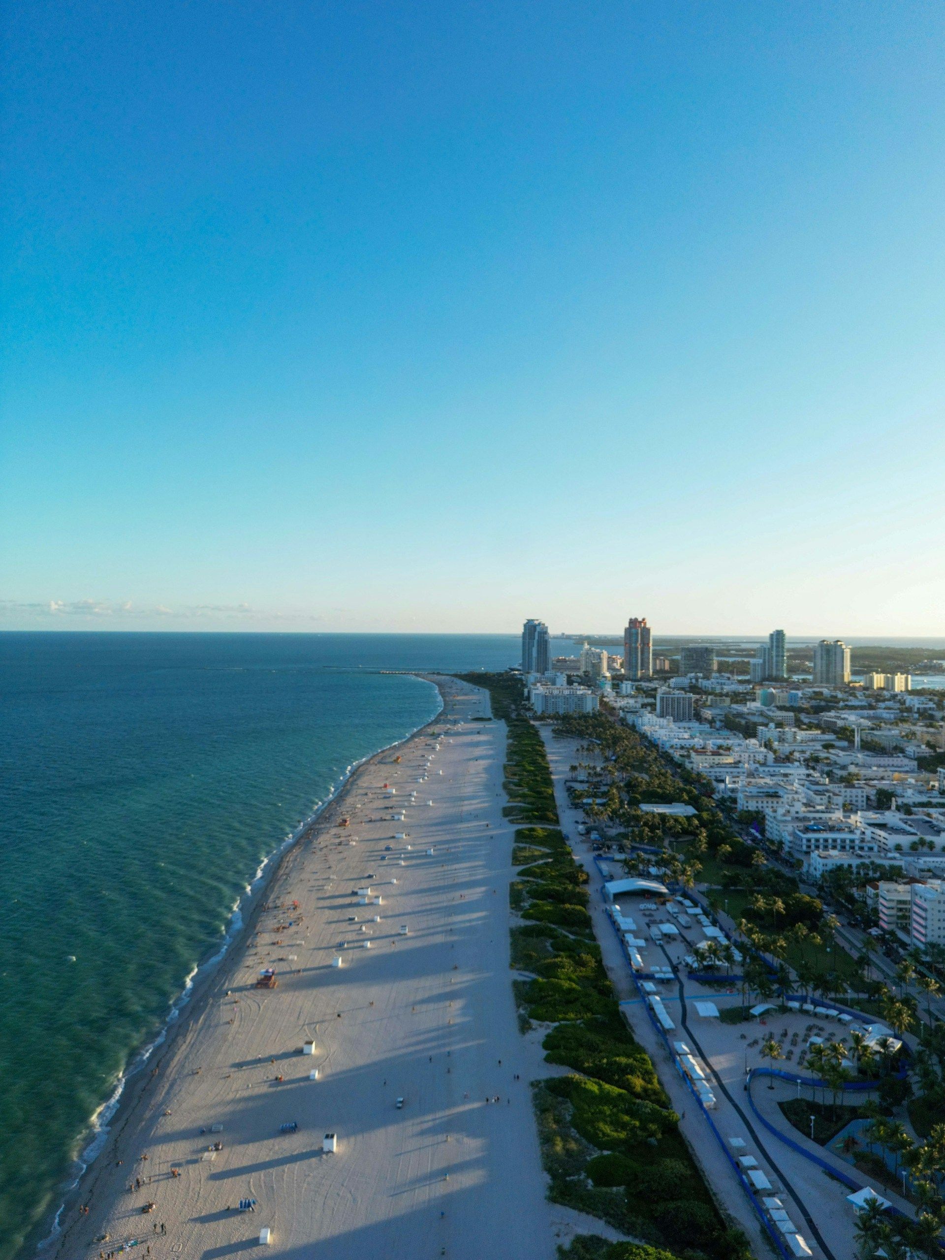 Aerial view of Miami Beach, Florida, with ocean on the left and buildings and sand on the right under a blue sky.