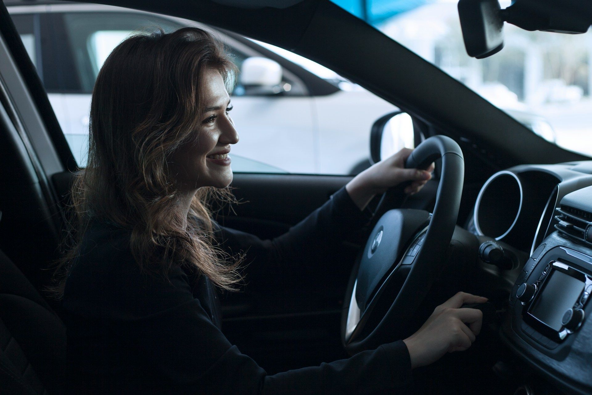 Woman smiles while driving a car, holding the steering wheel. Inside a car dealership.