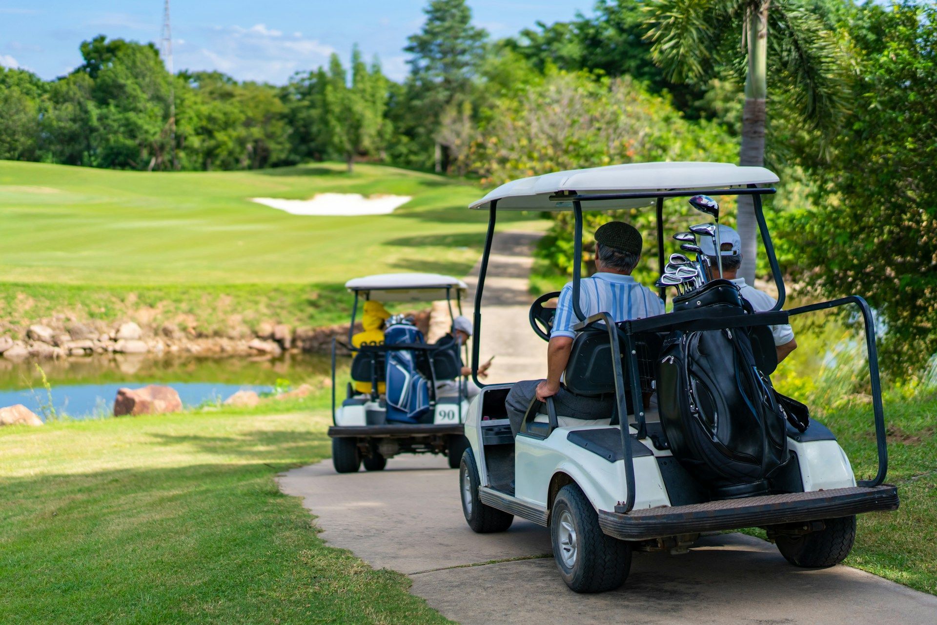 Two golf carts on a path beside a pond, heading towards a green on a golf course.