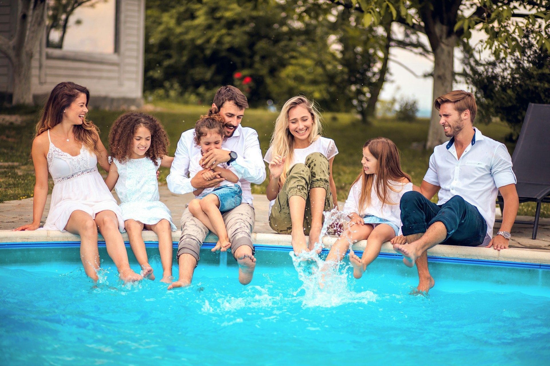 Family sitting on pool edge, splashing feet in blue water outdoors.