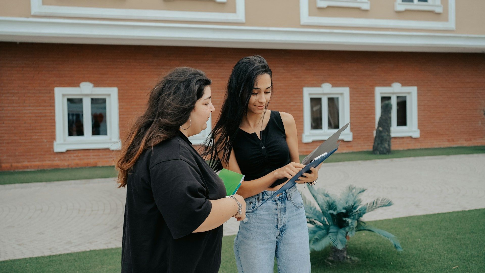 Two women reviewing a laptop outdoors near a brick building.