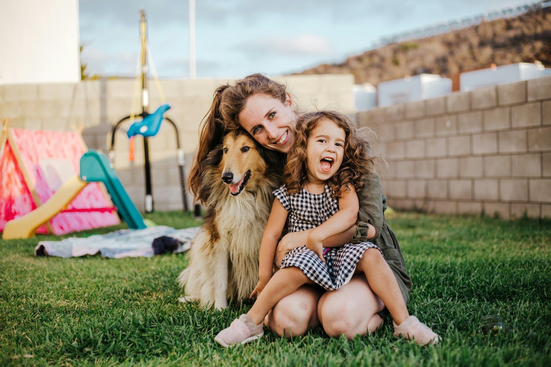 Woman and child sitting in grass with a dog, smiling. Child is laughing, and they are outside.