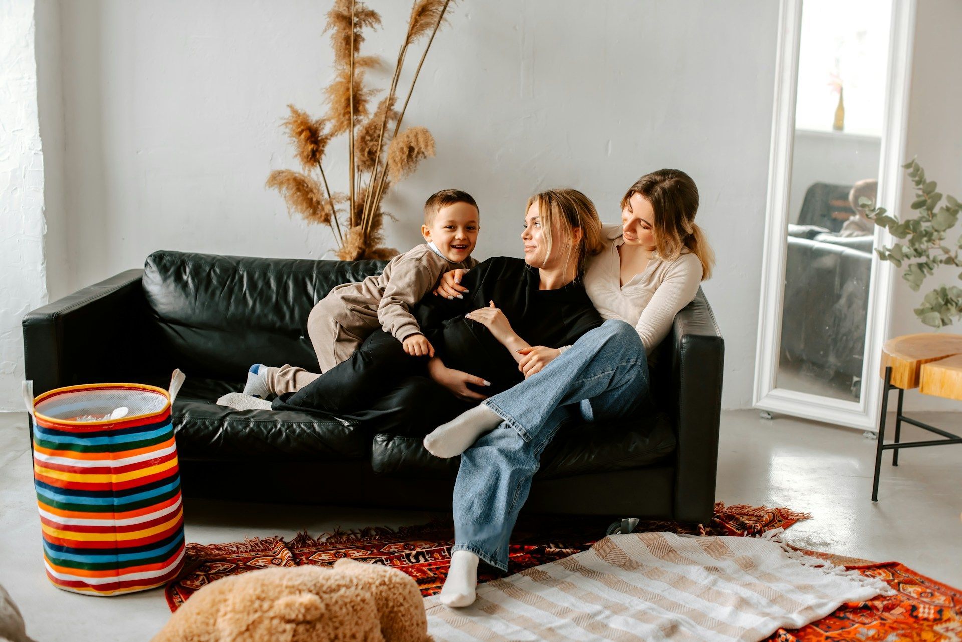 A pregnant person and two others sit on a black couch, laughing. A child playfully hugs the pregnant person.