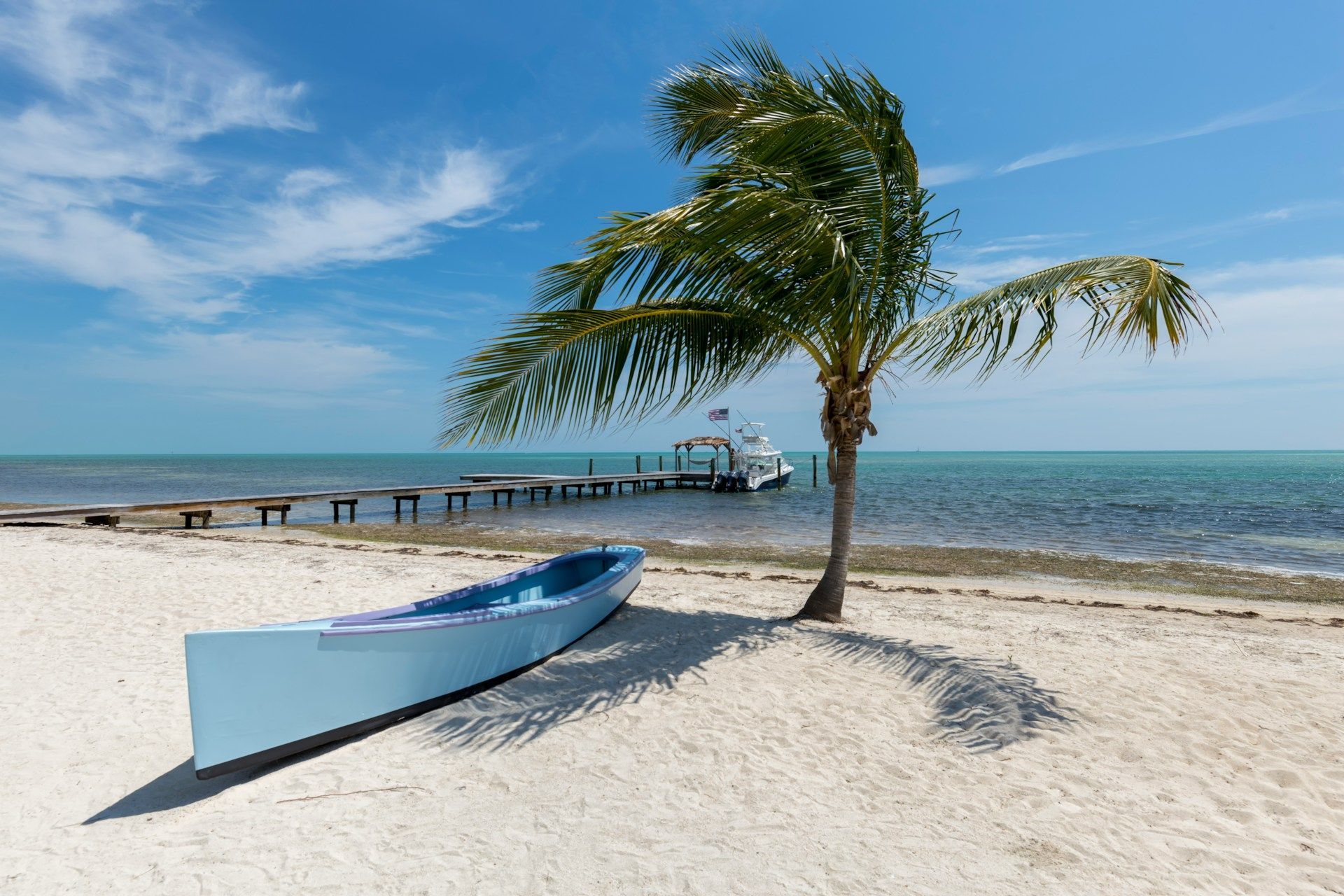 Blue boat on a white sandy beach with a palm tree, dock, and calm blue sea under a sunny sky.