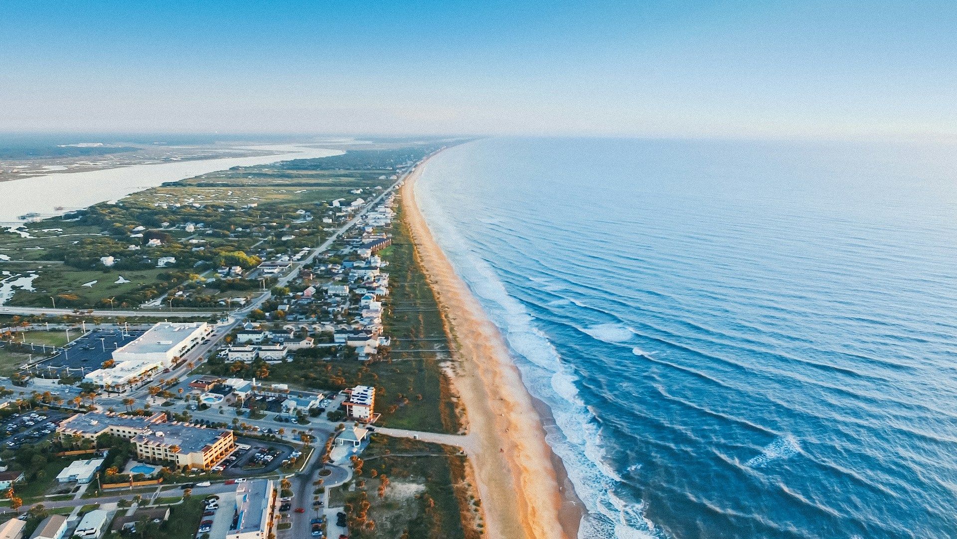 Aerial view of beach and coastline with buildings, sea, and river under a blue sky.