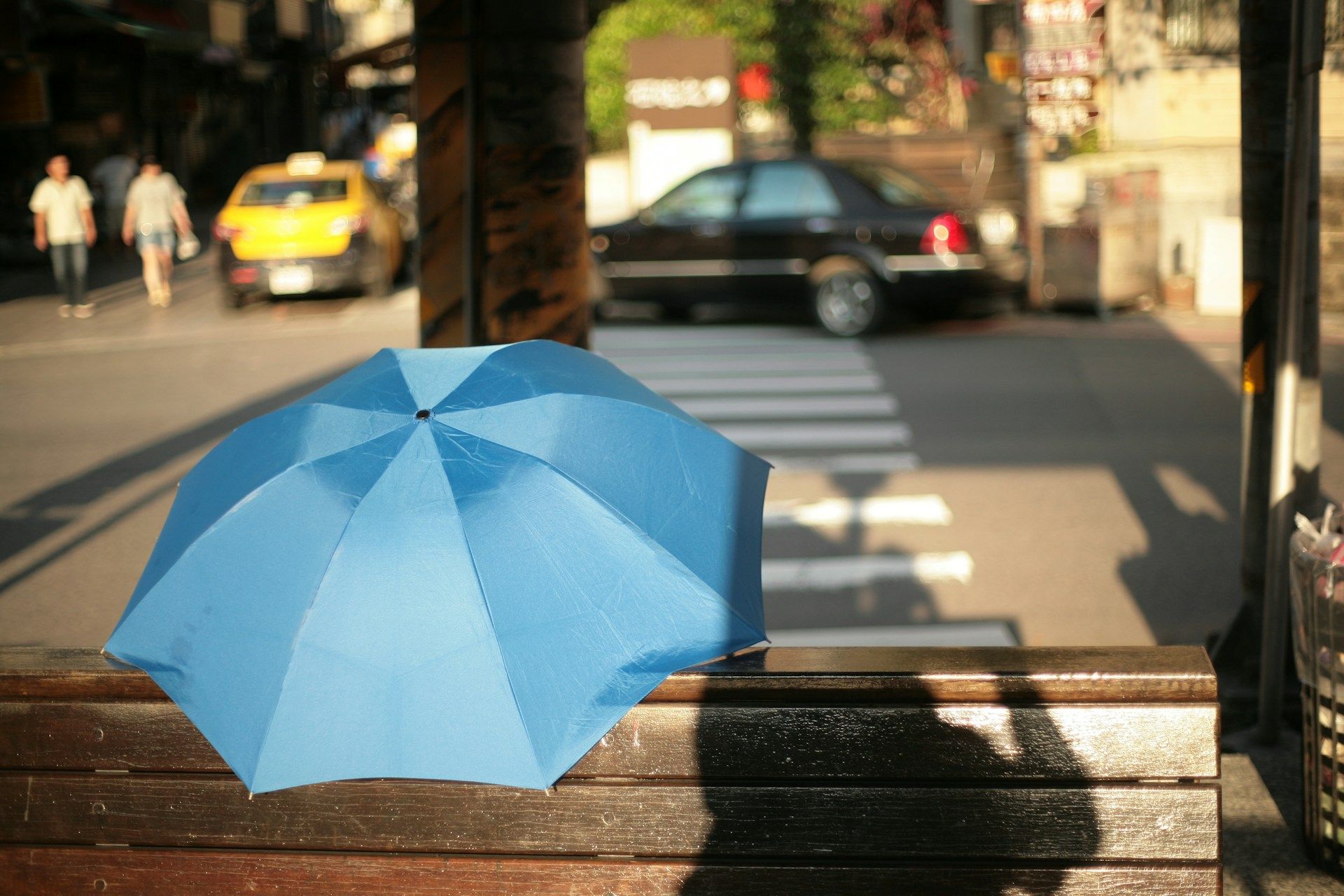 Blue umbrella on a wooden bench, street scene with pedestrian crossing, a taxi, and car.