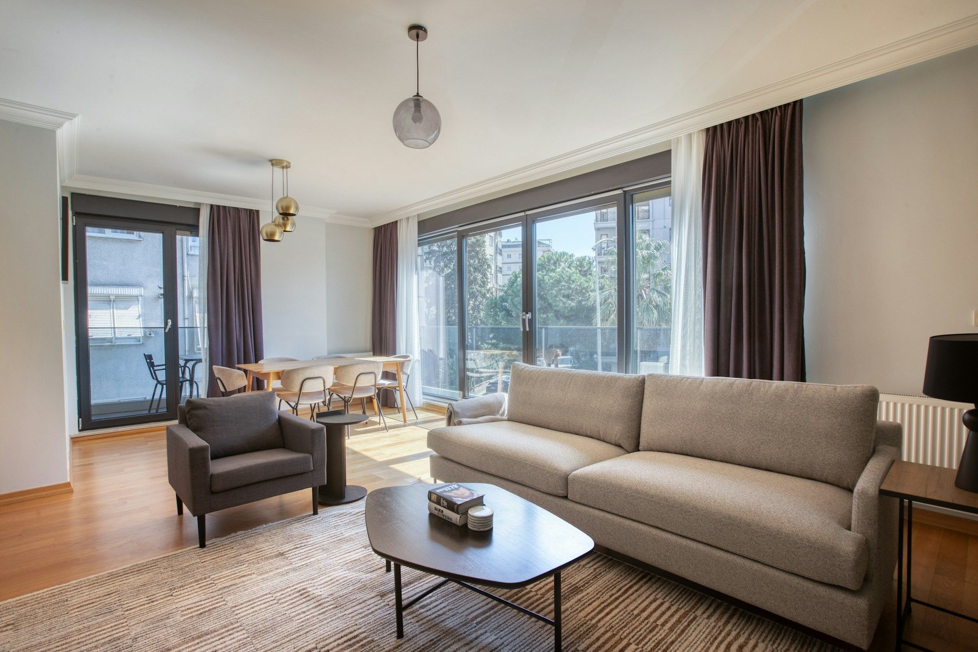 Spacious living room with sofa, armchair, coffee table, and large windows with curtains; dining area visible.