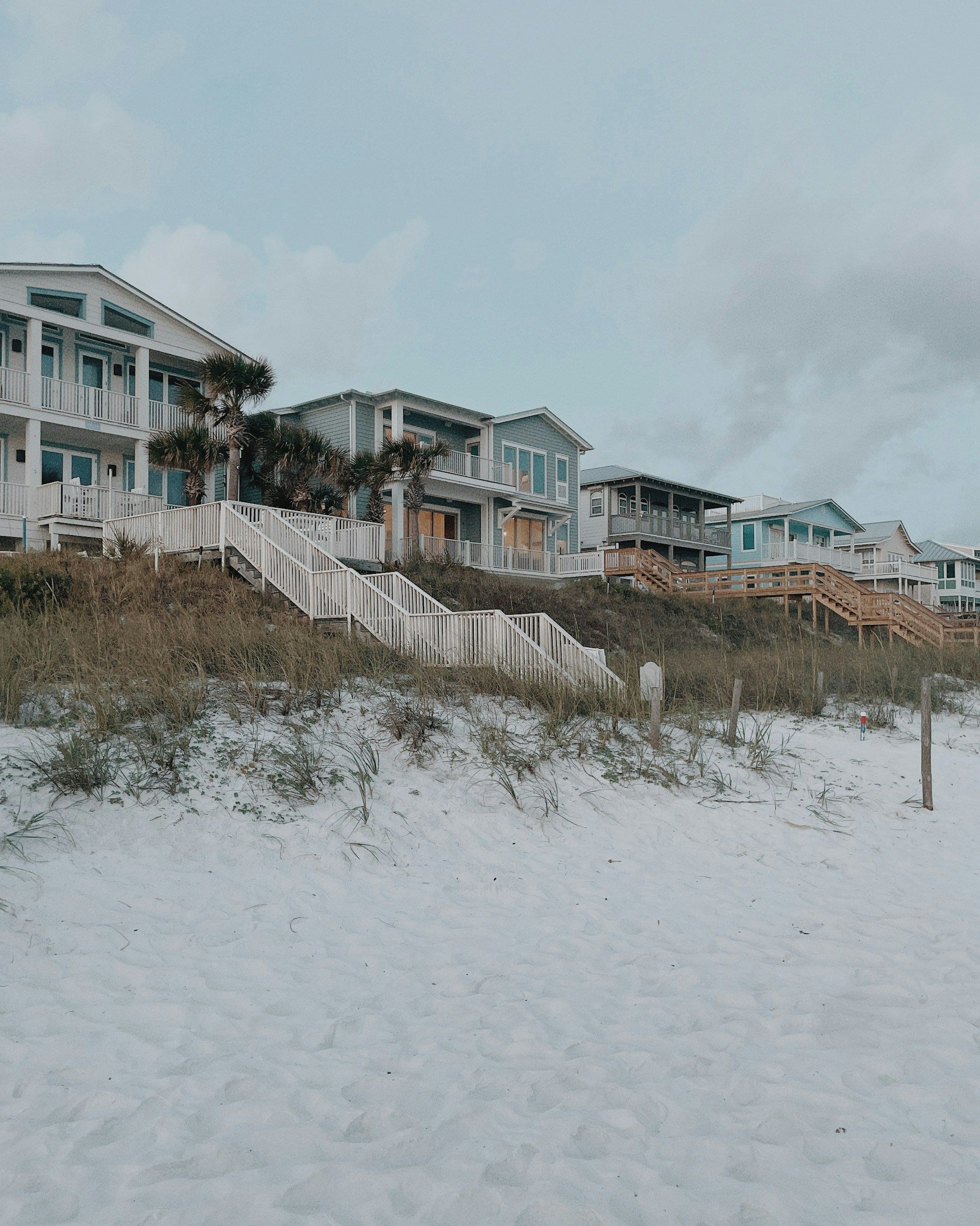 Beach houses line a sandy shore, white stairs leading to each, under a cloudy sky.