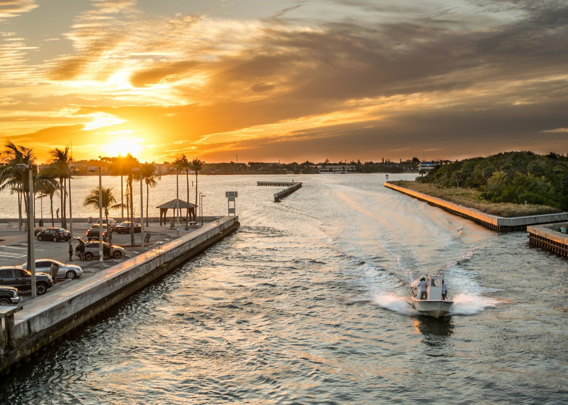 Sunset over a waterway; boat speeding toward the open ocean. Palm trees and buildings line the channel.