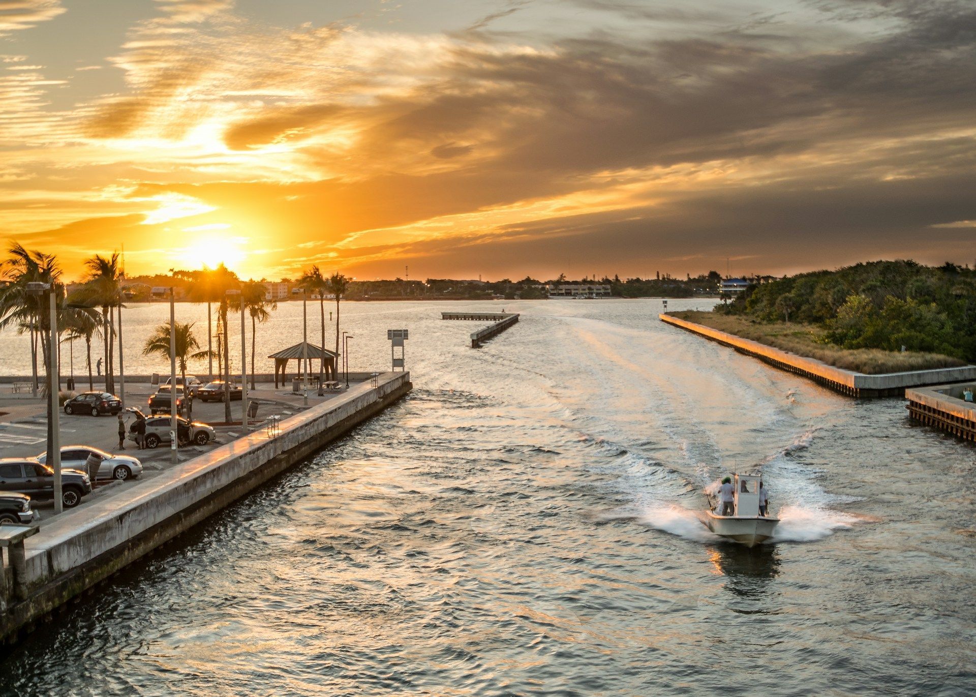 Sunset over waterway with boat, palm trees, and buildings, with cloudy sky.
