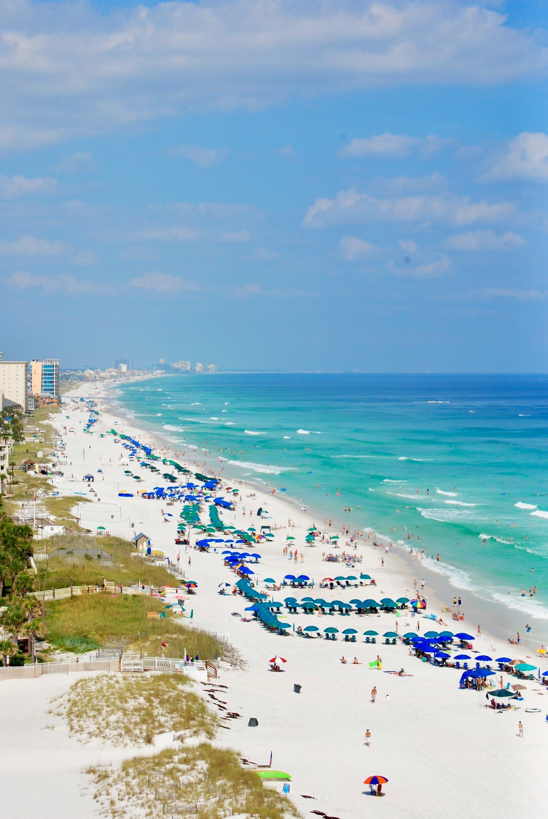 Aerial view of a white sandy beach, turquoise water, and a blue sky with clouds. Beachgoers with umbrellas.