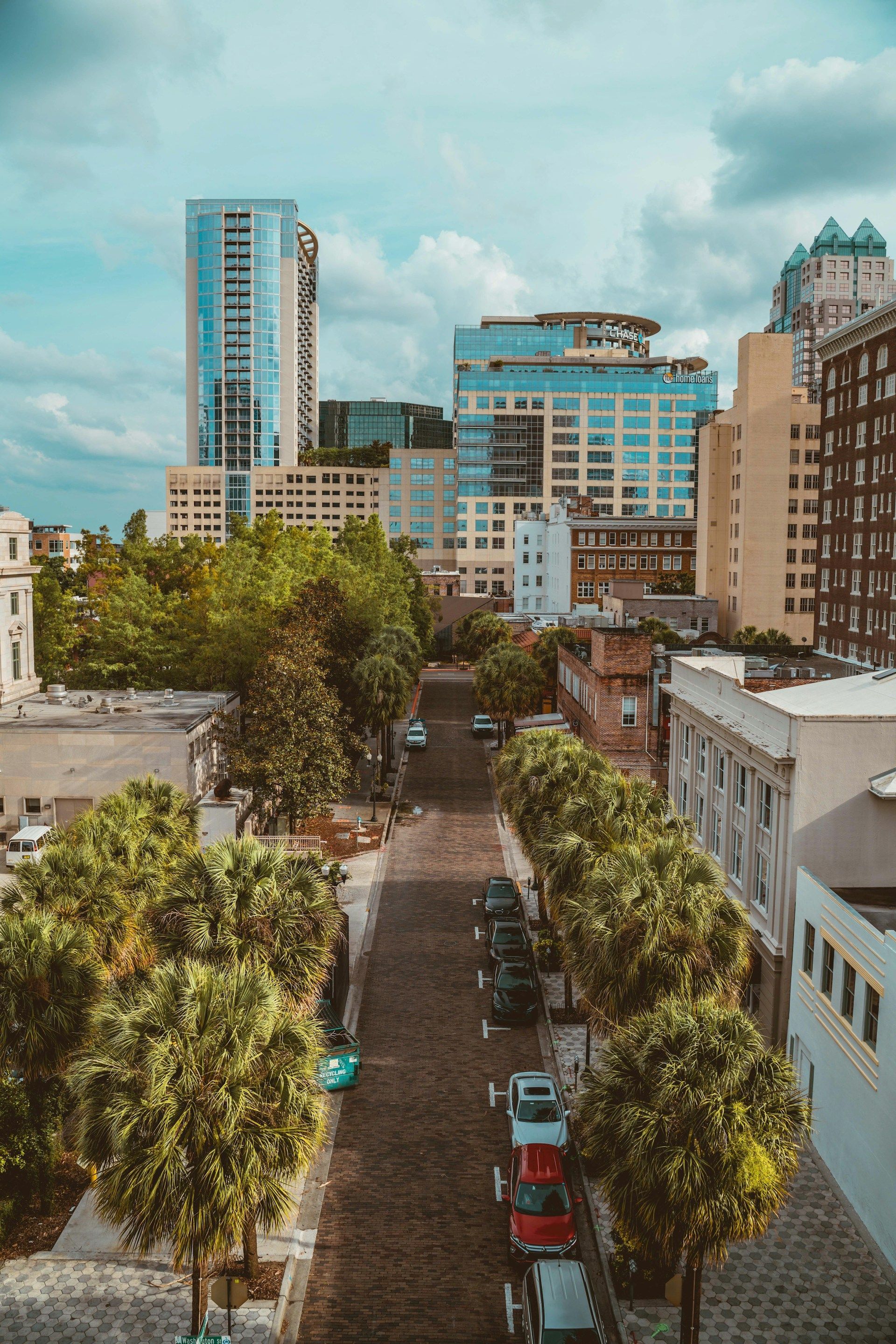 City street lined with trees and parked cars, tall buildings in the background under a cloudy sky.