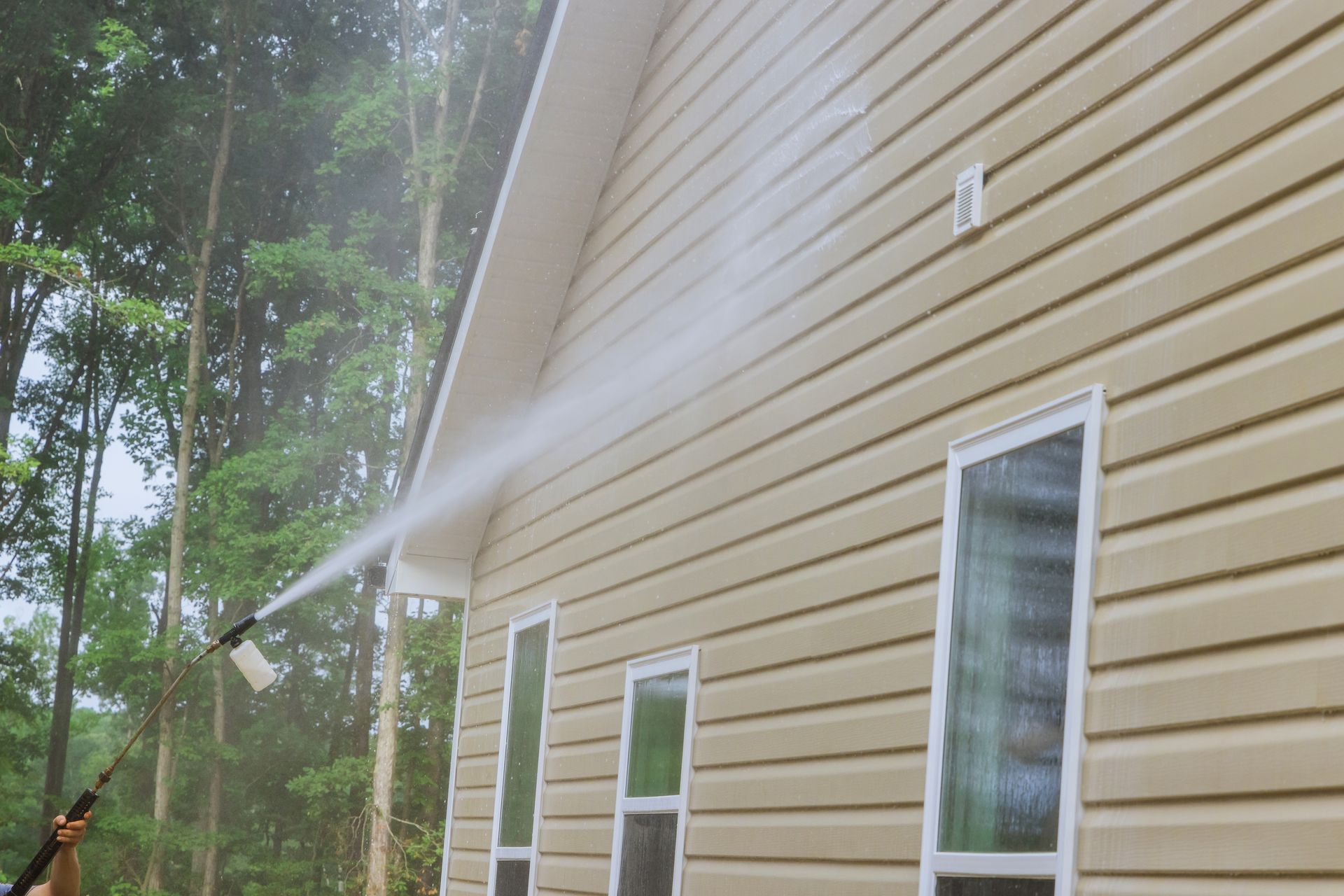 A person power washing the beige siding of a house, creating a stream of water. The house has white trim and windows.