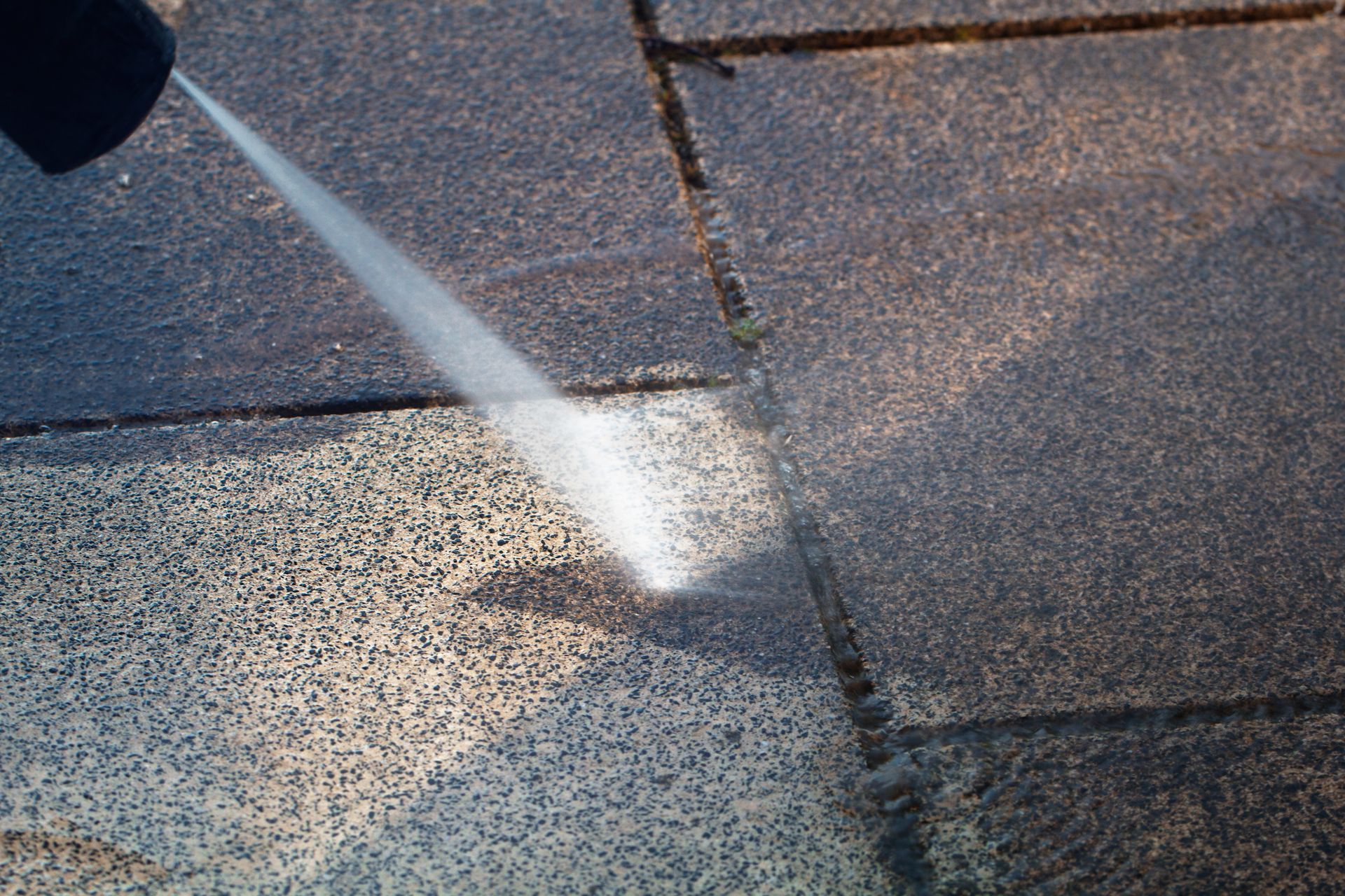 Power washer cleaning a dirty, dark gray brick patio, showing a visible contrast between the cleaned and uncleaned areas.