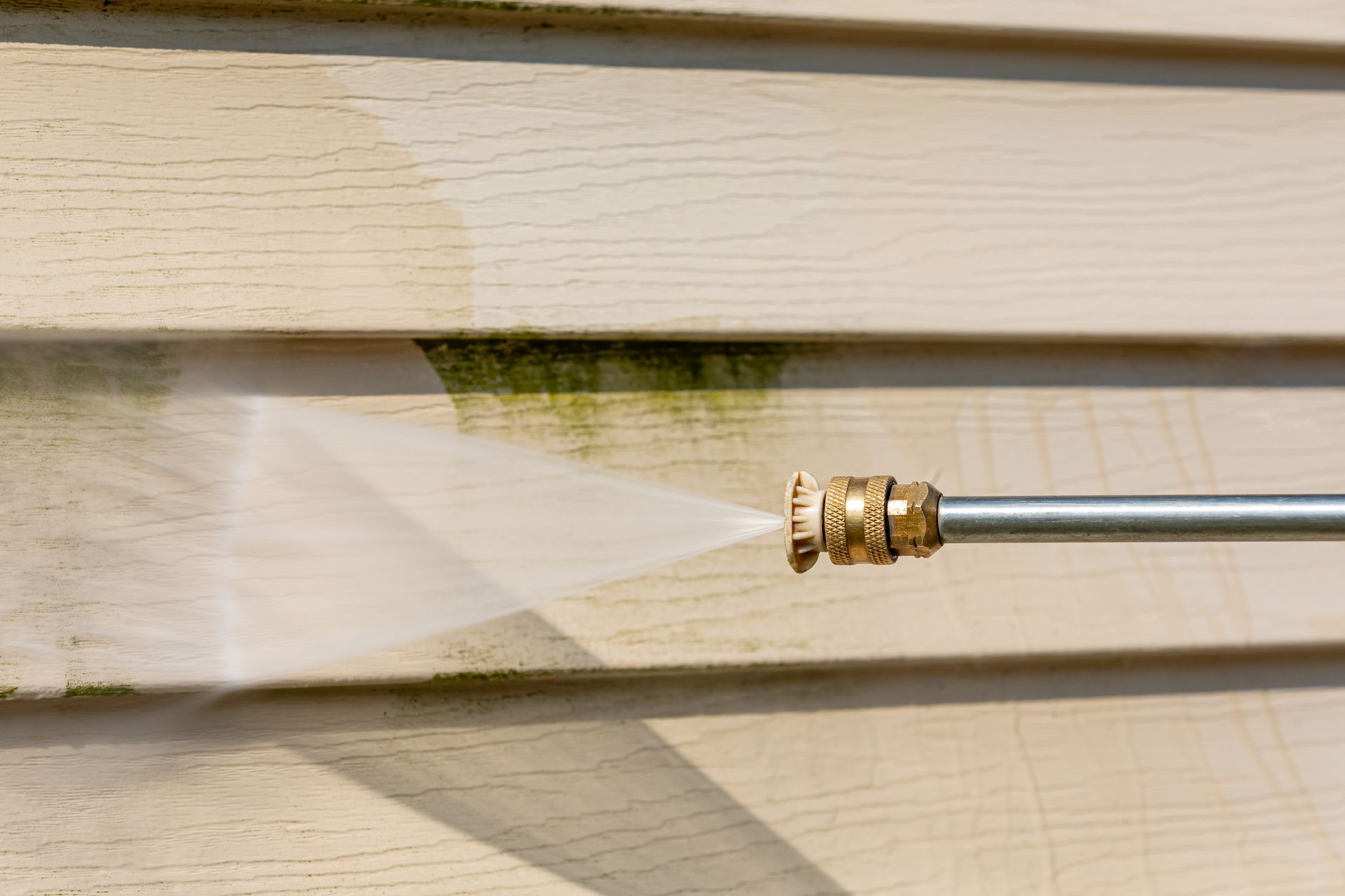 Pressure washer spraying a beige vinyl siding, removing green algae and dirt.