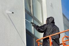 Person in black waterproof gear power washing a building facade with windows from a raised platform.