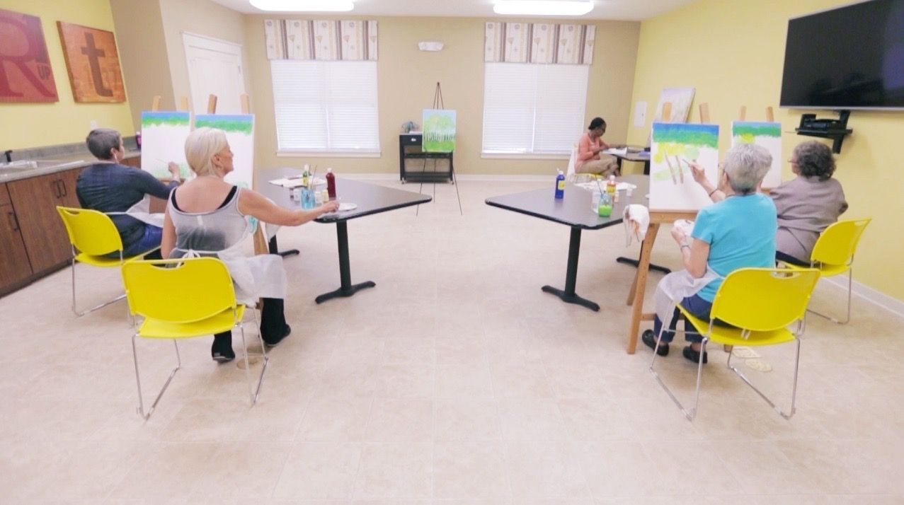 Residents sit on bright yellow chairs painting canvases in a sunny communal art room.