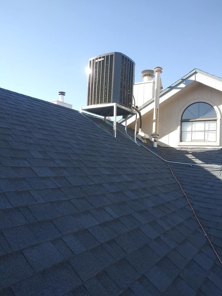 An air conditioning unit sits on a metal stand atop a dark shingled roof, with chimney in background.