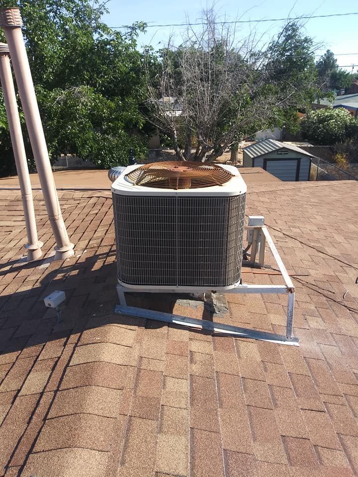 Air conditioner unit on a brown shingled roof with trees in the background.