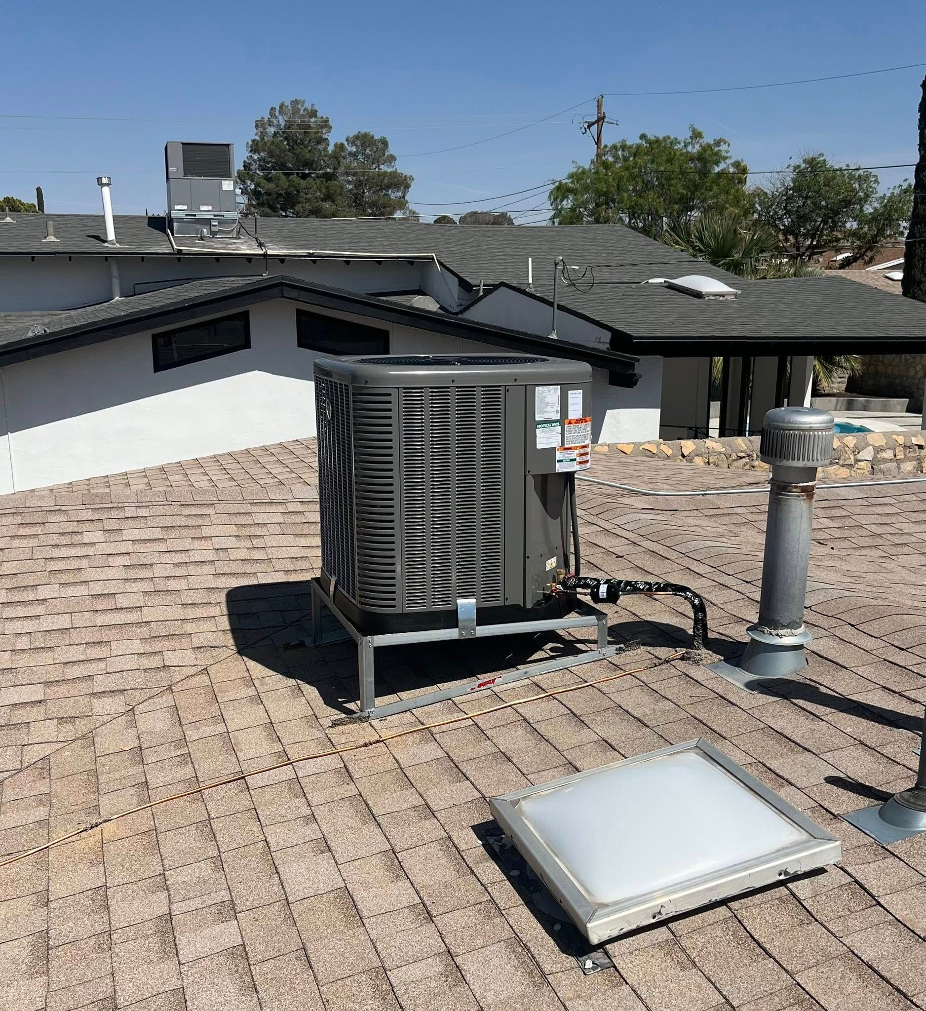 Air conditioning unit on a brown shingle roof, near a skylight and vent against a sunny sky.