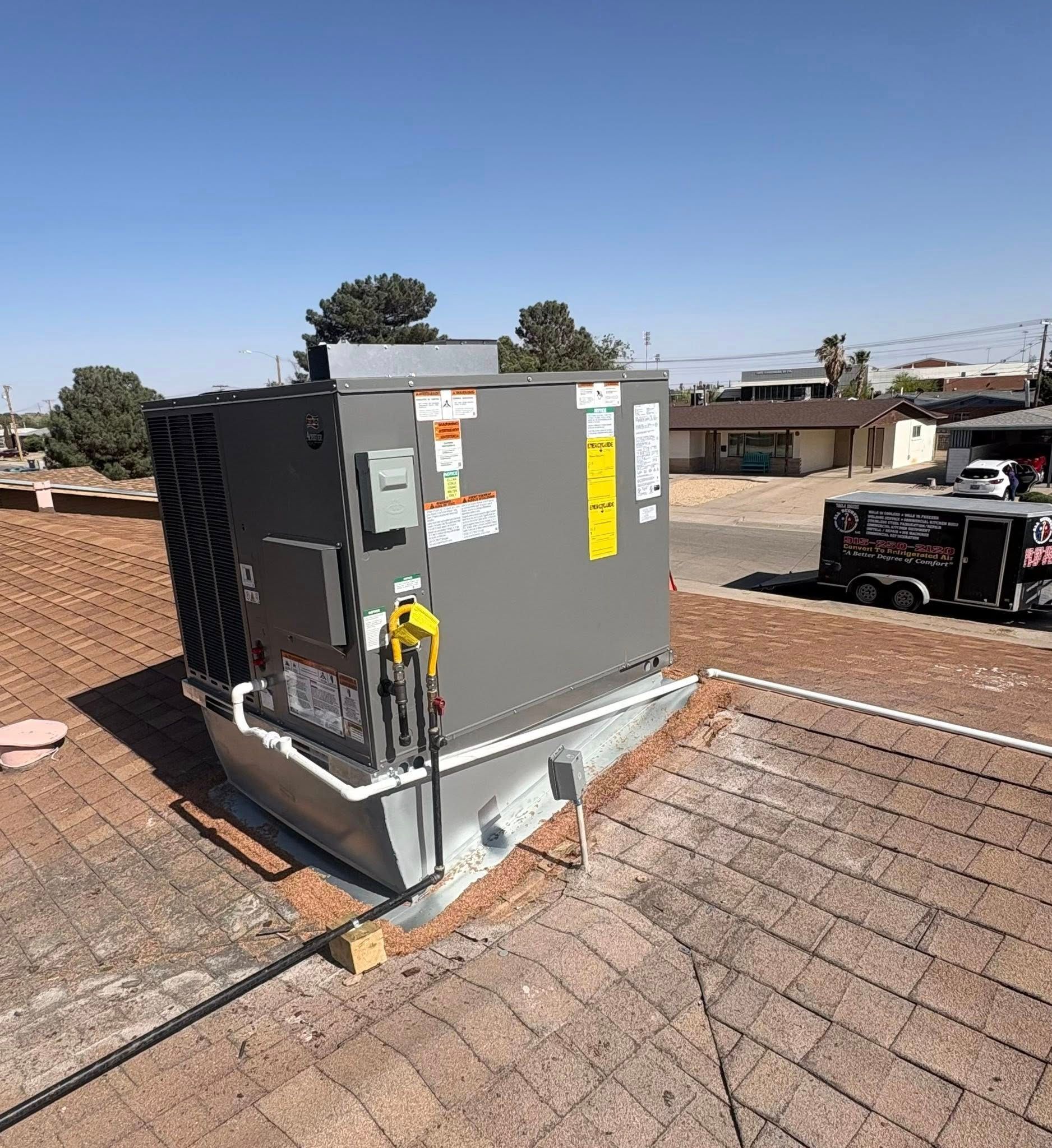 HVAC unit installed on a rooftop with clear sky. Grey unit, white pipes, brown roof. A trailer is parked nearby.