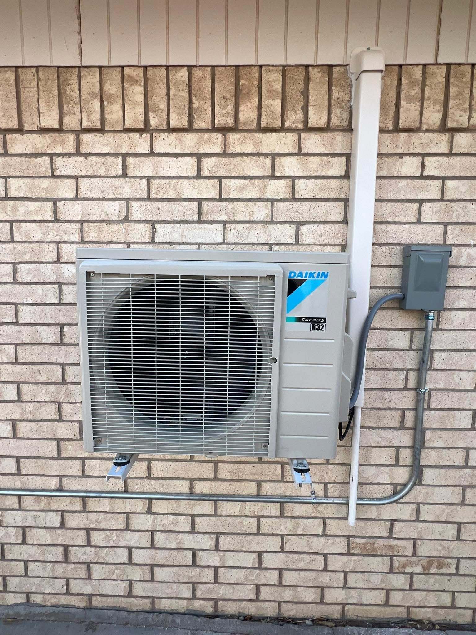 Wall-mounted air conditioner unit with a brick wall background, electrical components visible.