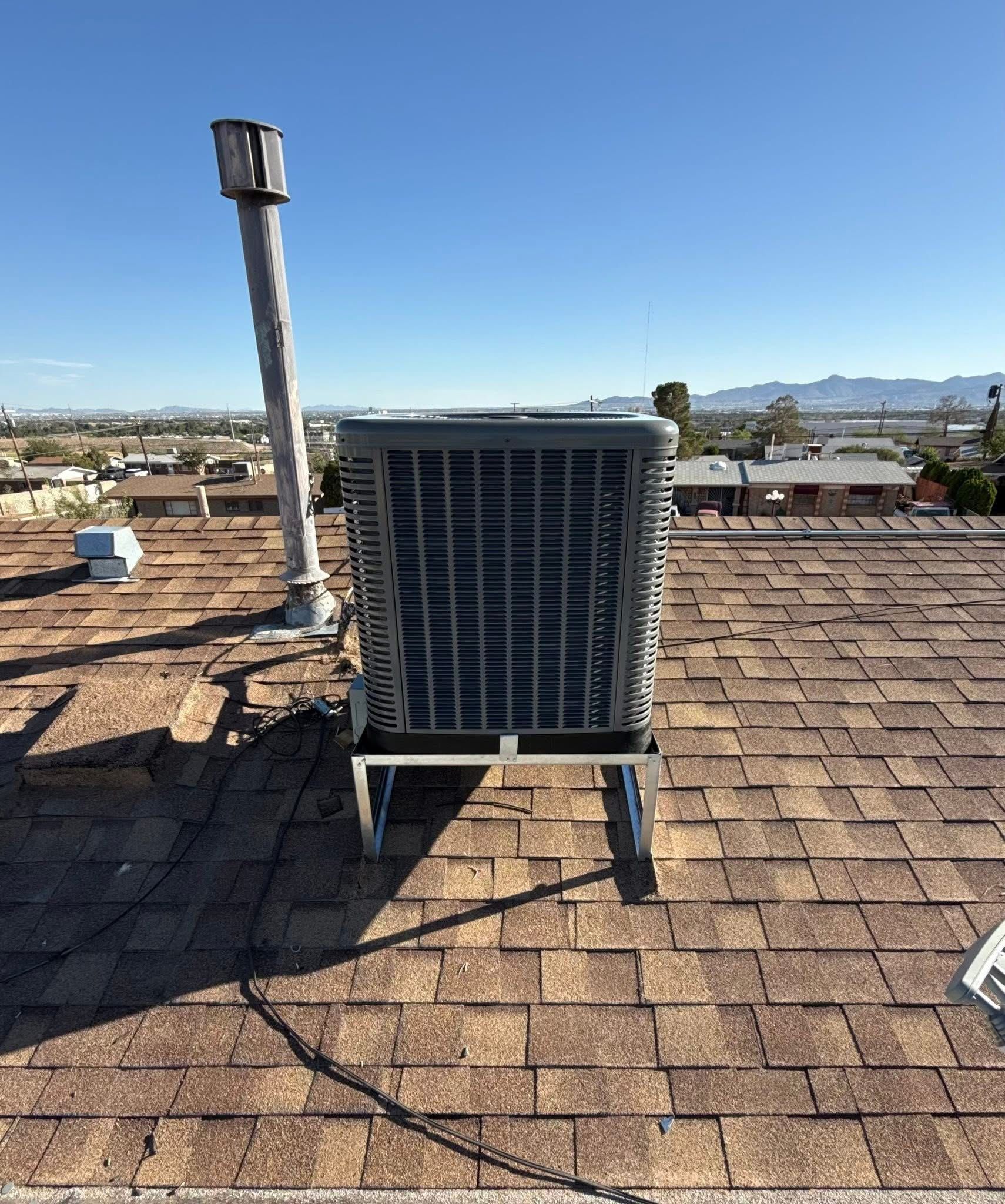 An air conditioning unit on a brown shingle roof with a chimney and a clear blue sky background.