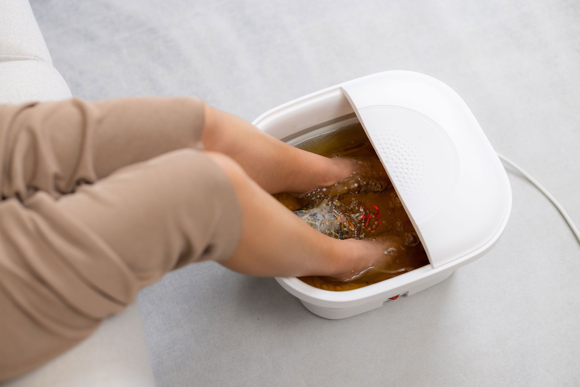 Feet soaking in a white foot spa filled with brown liquid, light tan pants visible.