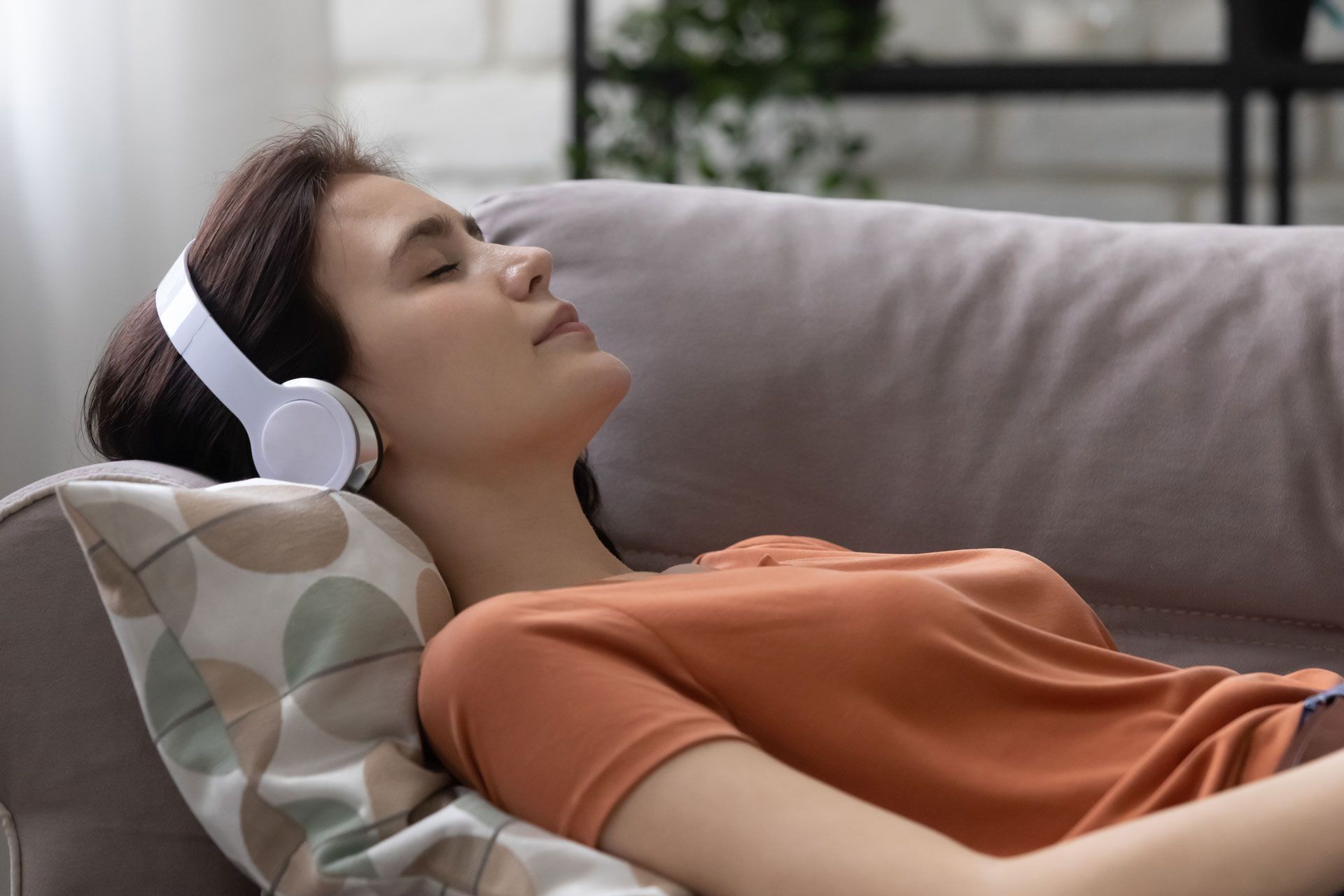 Woman wearing headphones reclines on a couch with eyes closed, appearing relaxed.