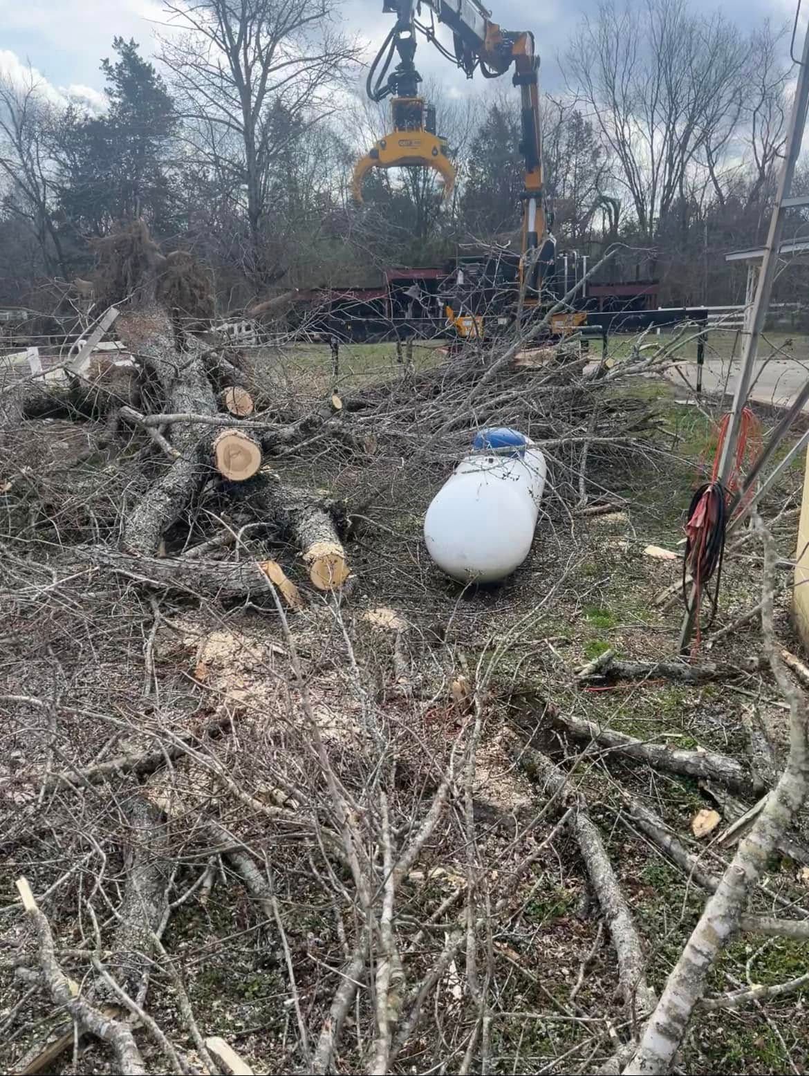 A crane lifts a yellow bucket over a brush pile beside a white propane tank in a wooded clearing.