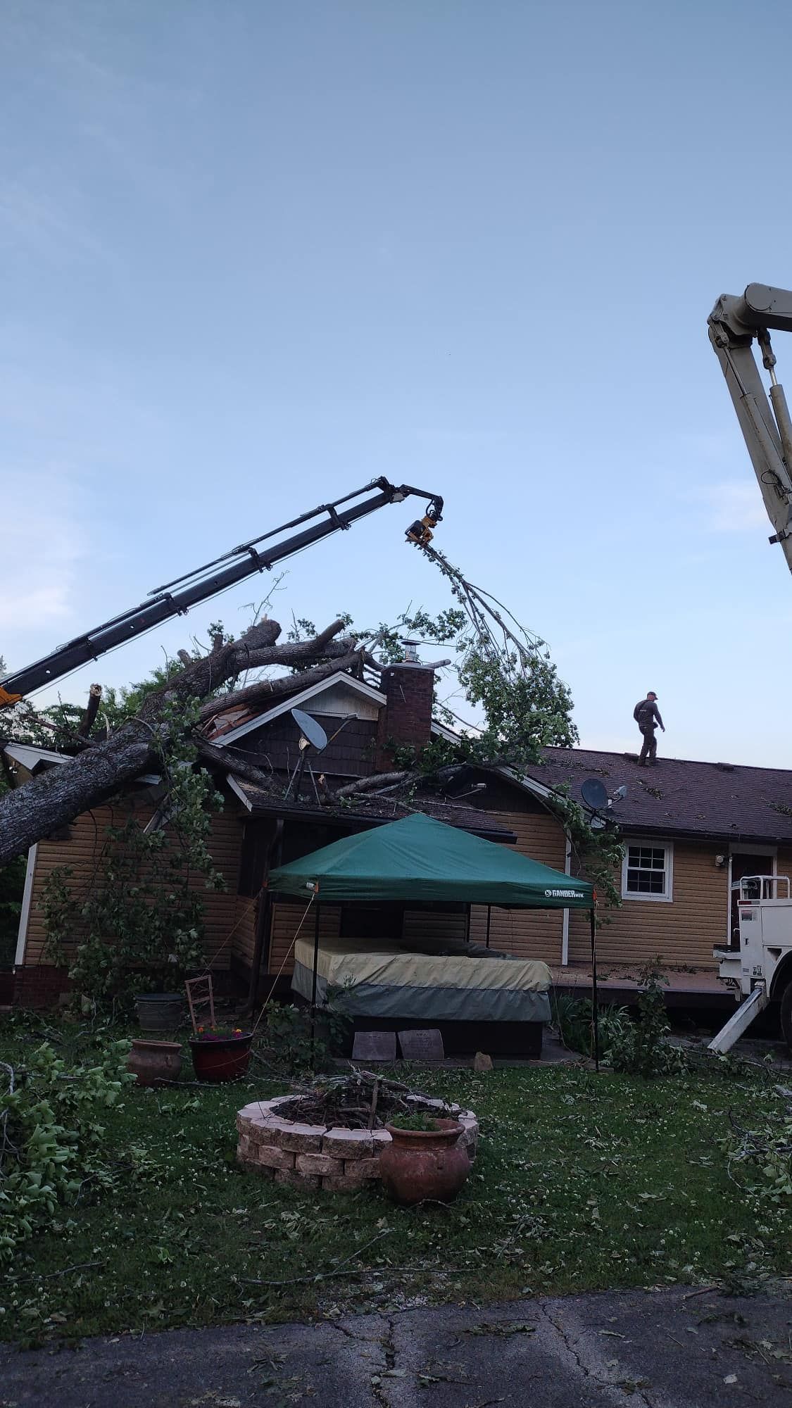 Collapsed house with a damaged roof, two crane arms, and a worker standing on the debris in a yard.
