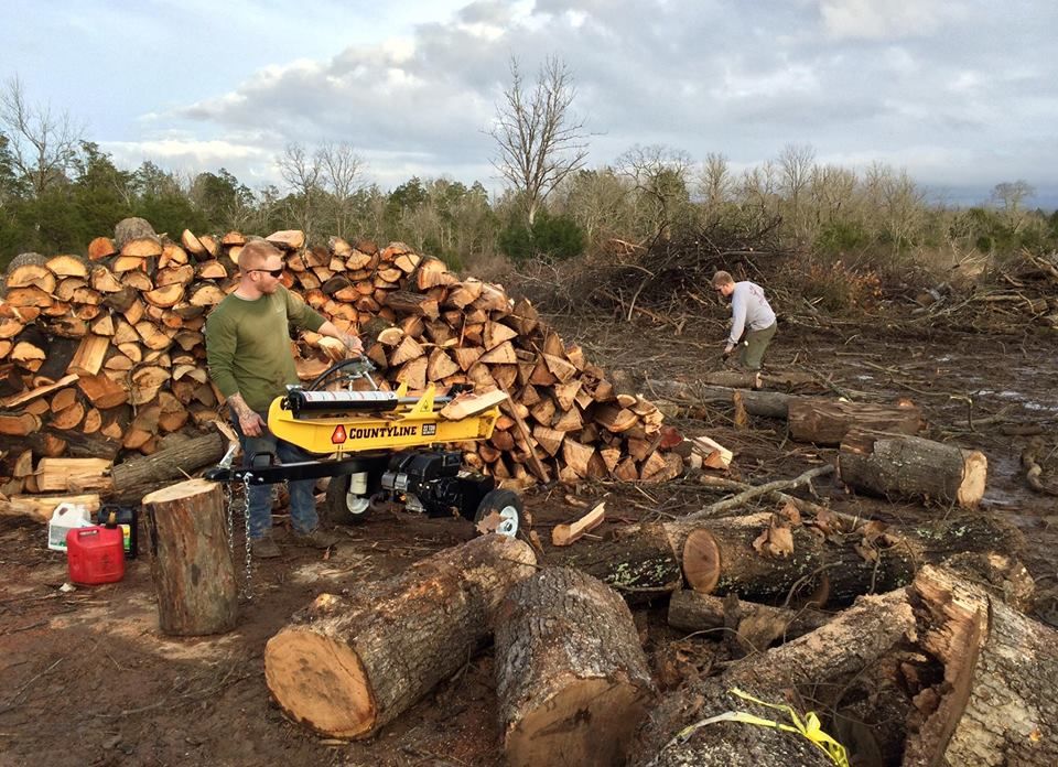 Two people working among stacks of cut logs and a yellow wood splitter in a wooded clearing