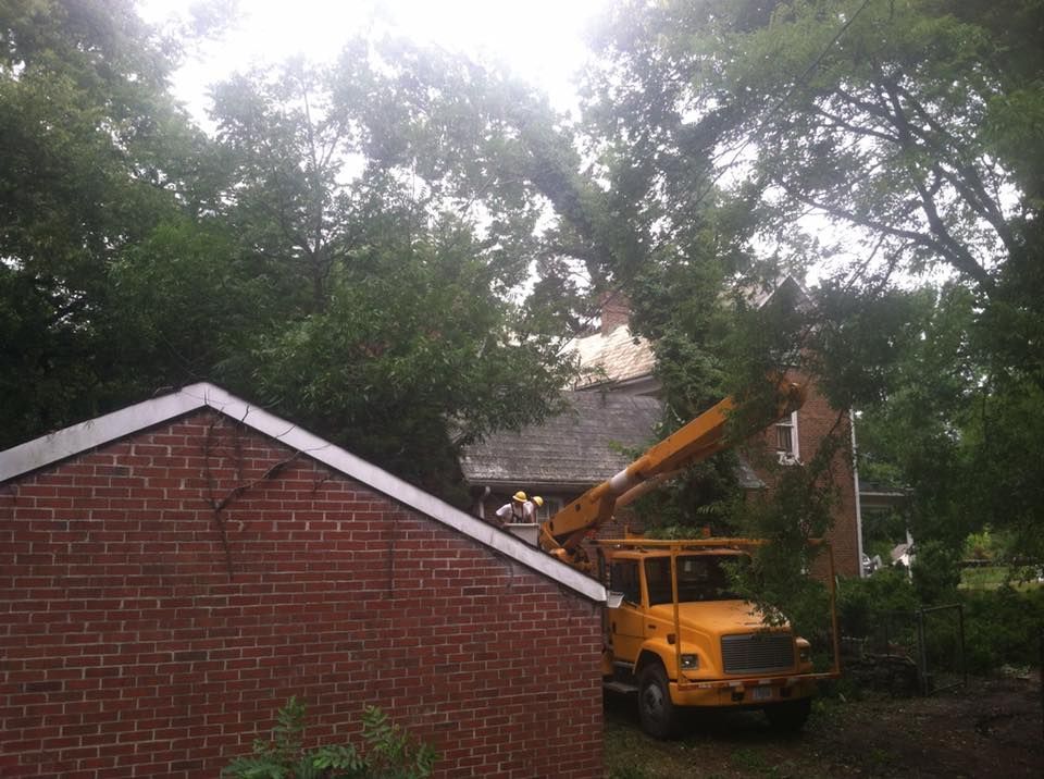 Yellow utility truck with raised boom working beside a brick house under trees