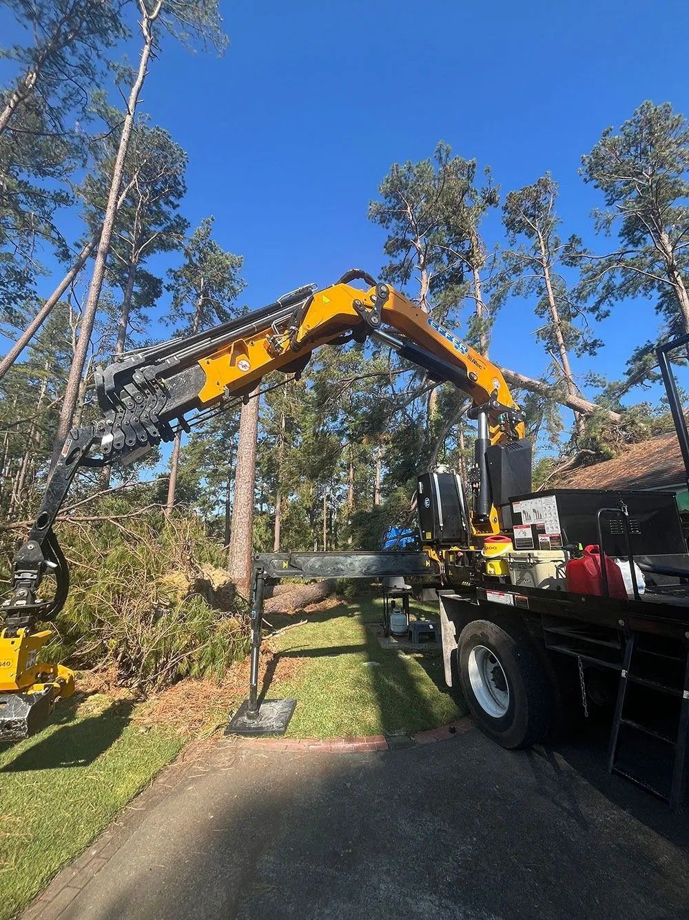 Orange crane truck lifting a boom arm in a wooded yard under a clear blue sky