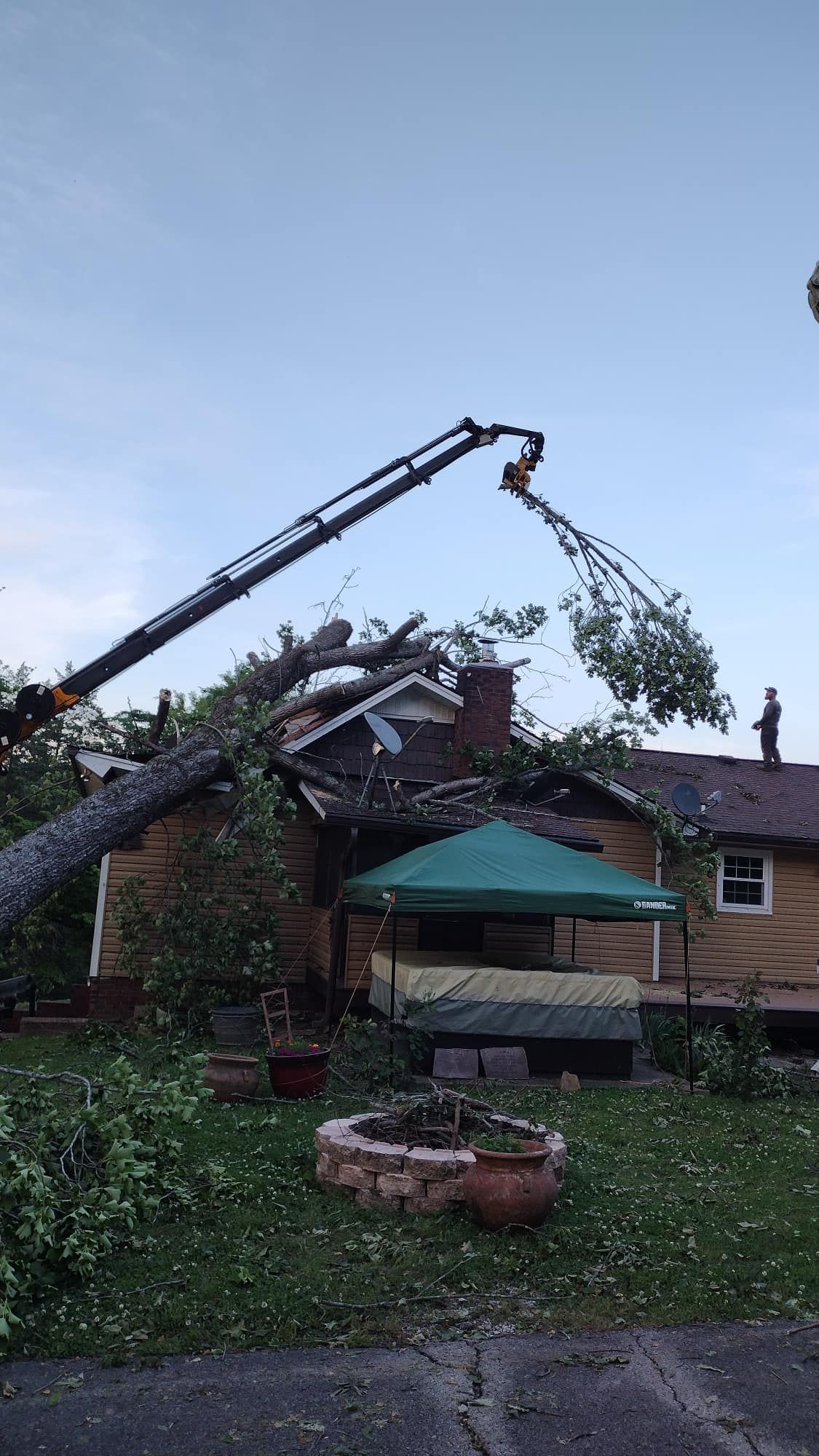 Crane lifting storm-damaged roof debris from a house in a suburban yard at dusk