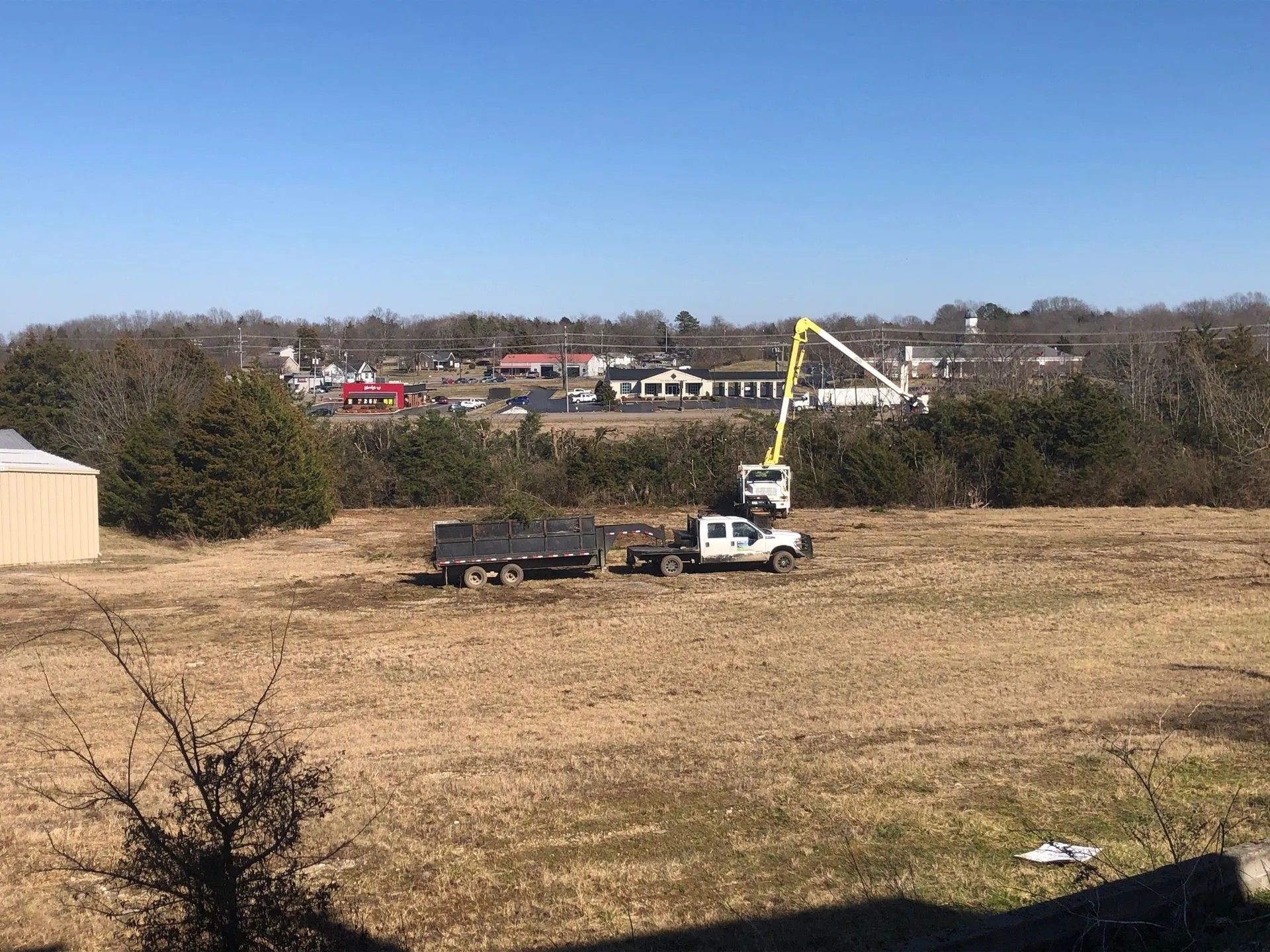 White utility truck with boom lift parked in a field near houses and trees under a clear sky