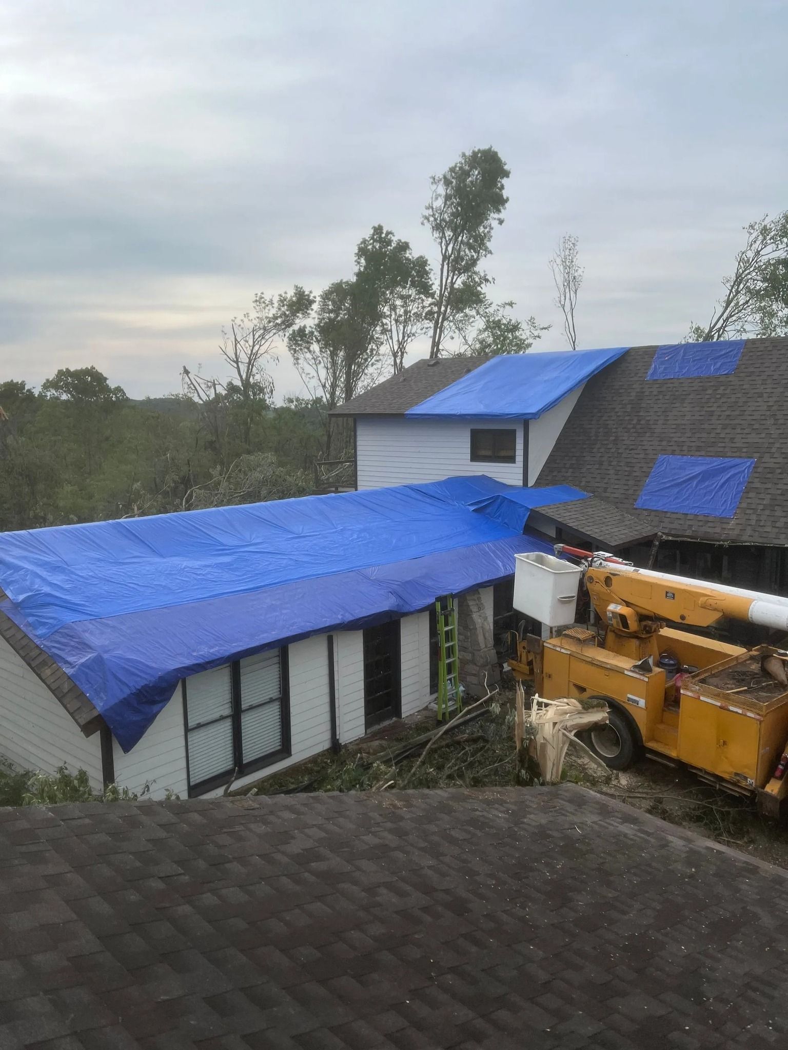 Damaged building with blue tarp roof beside a yellow excavator in a muddy area under cloudy sky