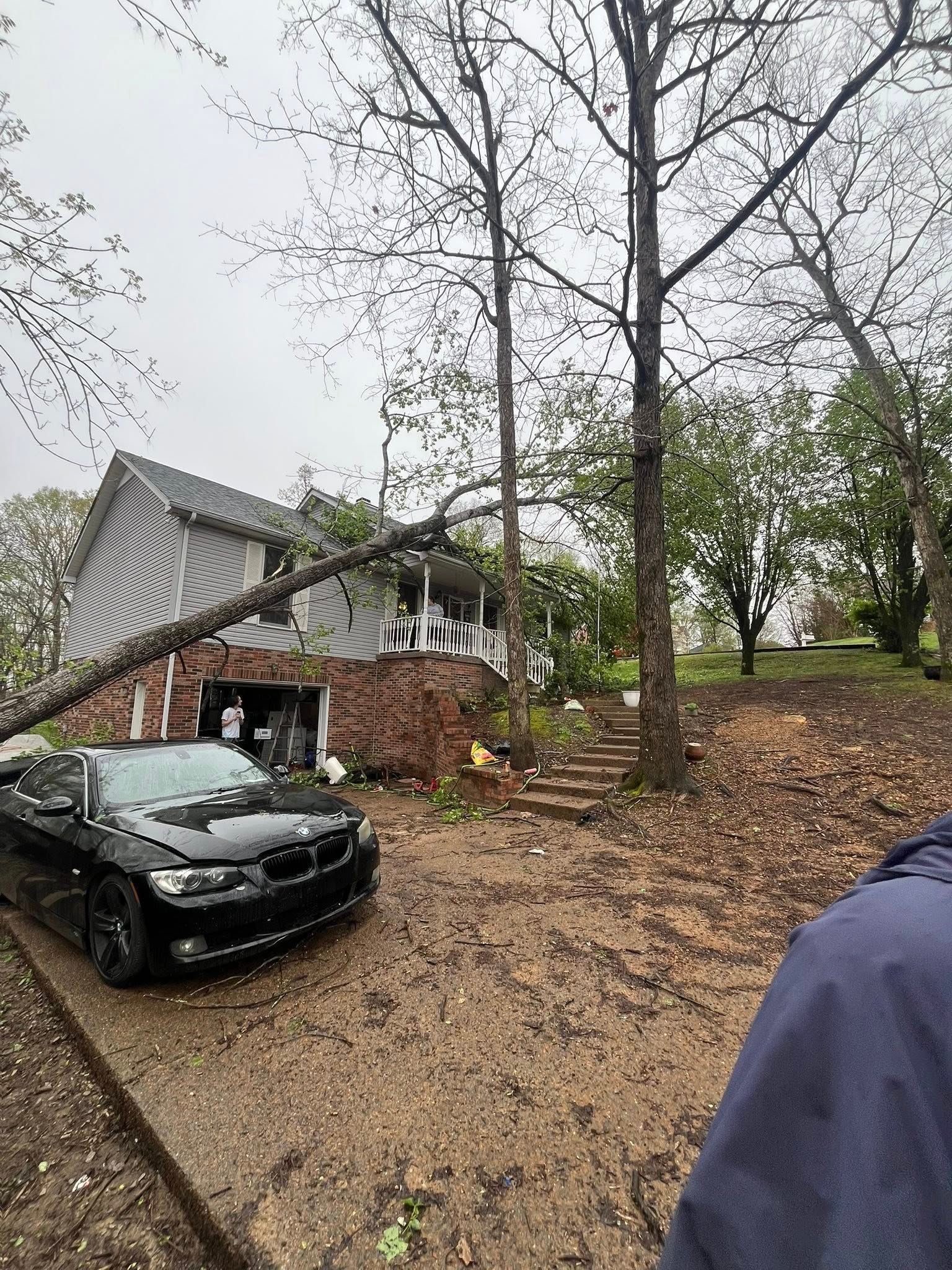 Black car near a damaged house and fallen tree after a storm, with debris on the ground.