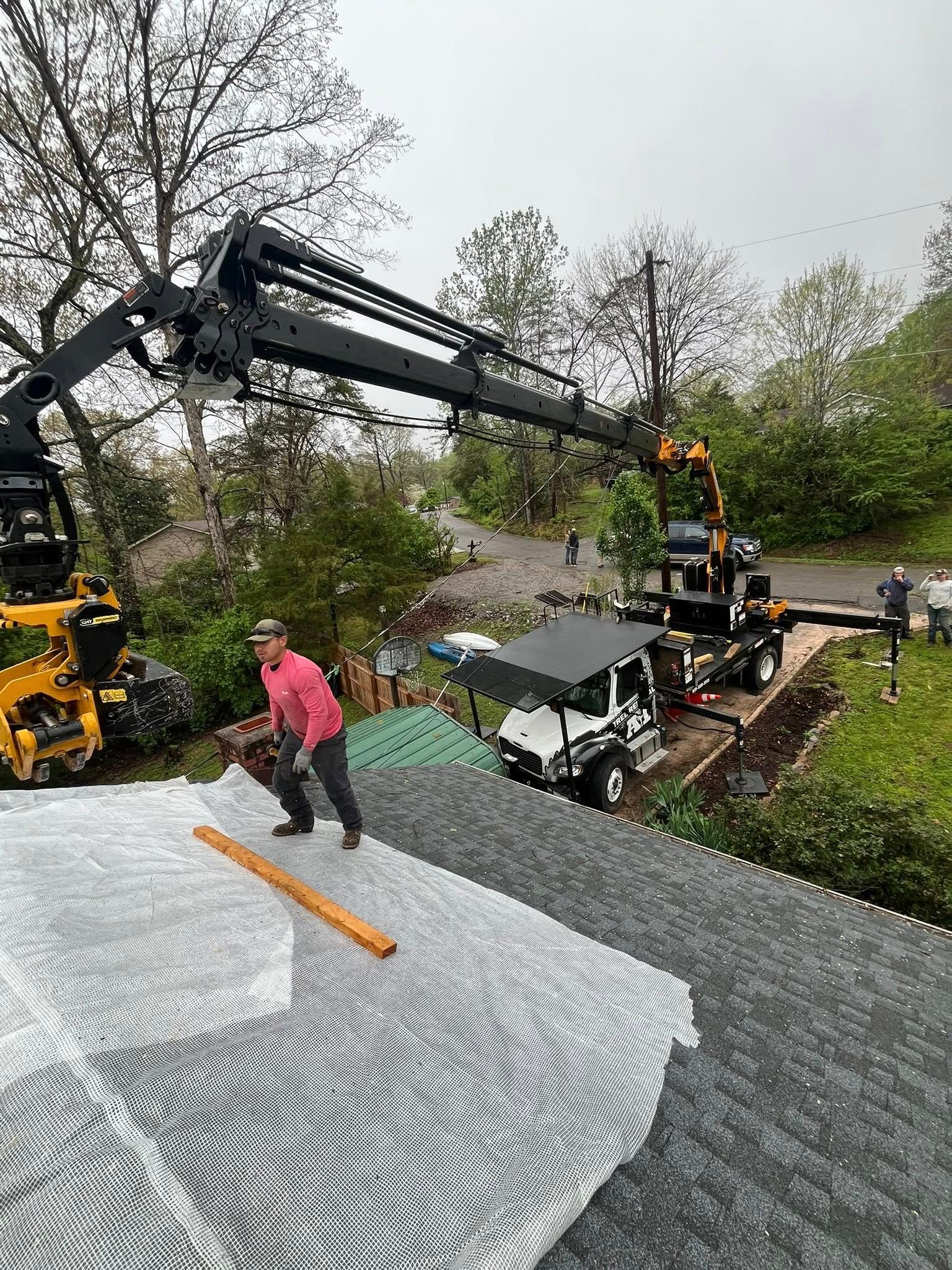 Workers use a crane to lift roofing materials beside a house in a rainy yard