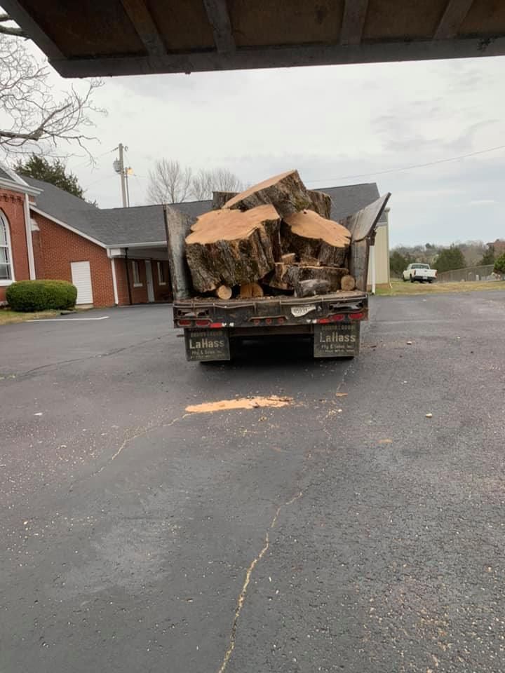 Truck loaded with cut firewood parked in a residential driveway, with wood chips scattered on the pavement.