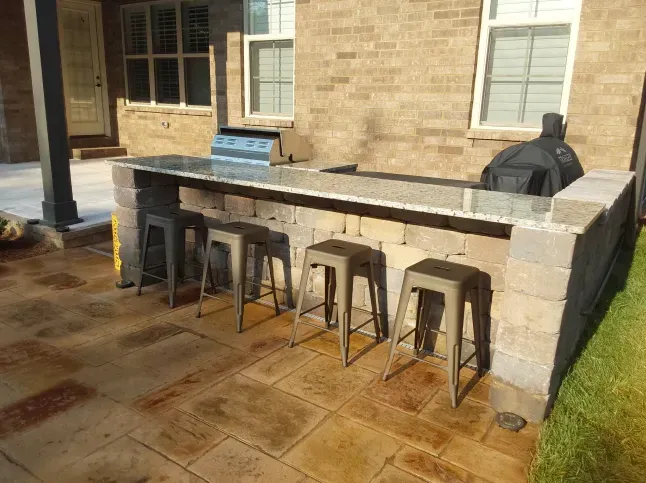 A patio with a grill and stools in front of a brick house.