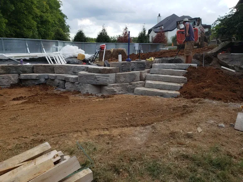 A man is standing in the dirt next to a pile of wood.