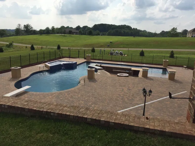 A large swimming pool is surrounded by a brick patio and a fence.