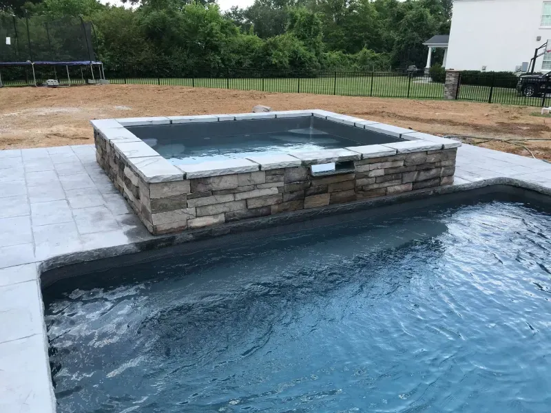 A square swimming pool with a stone wall and a trampoline in the background.