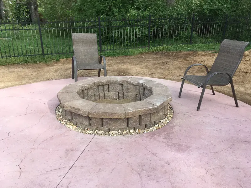 A fire pit is sitting on top of a concrete patio next to two chairs.