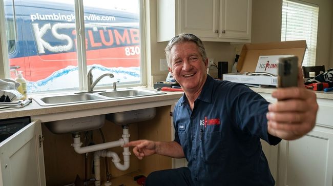 Plumber smiling in a kitchen, holding a tool and pointing under the sink next to a window.
